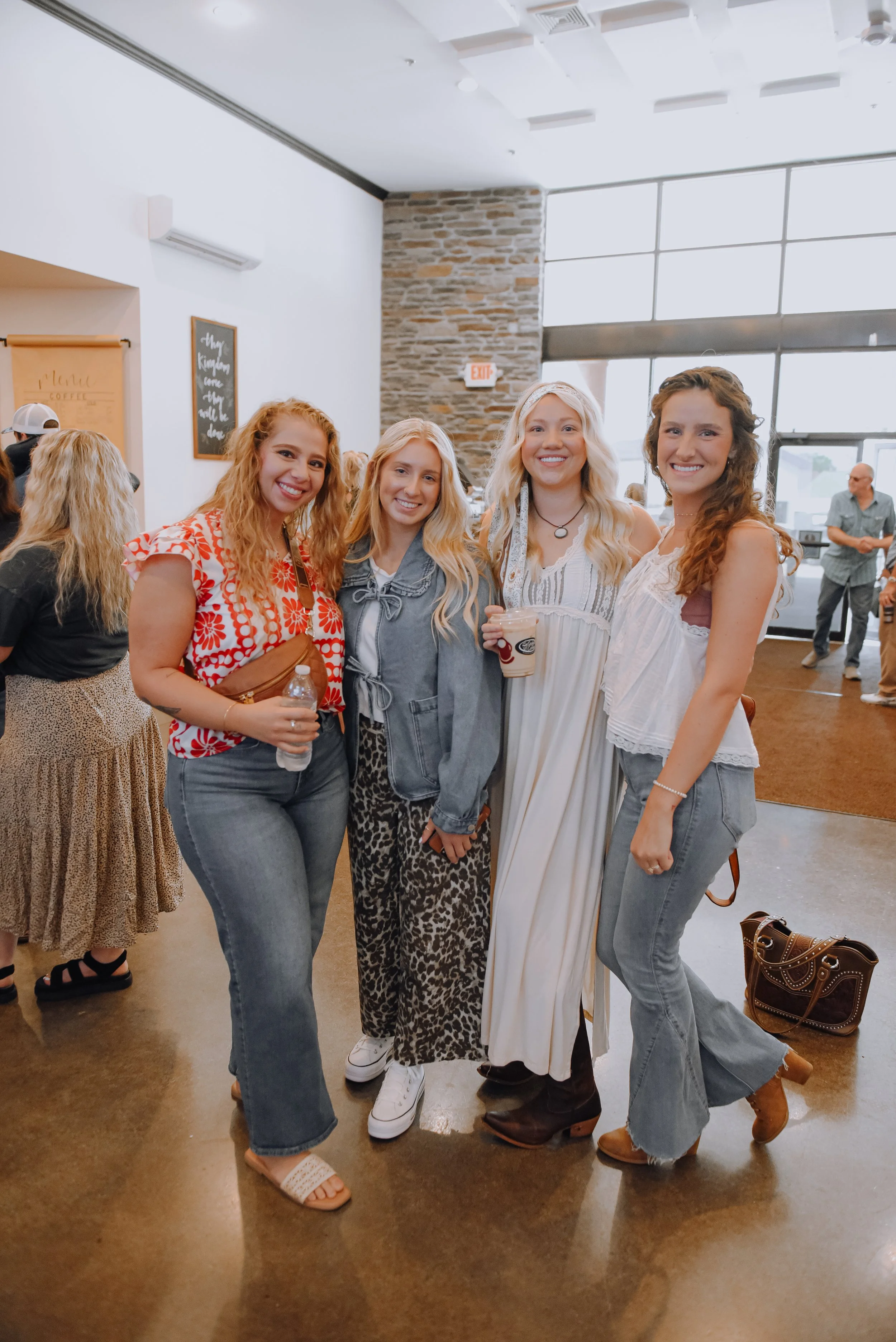 Four women standing together indoors, smiling at the camera, with other people and a brick wall in the background.