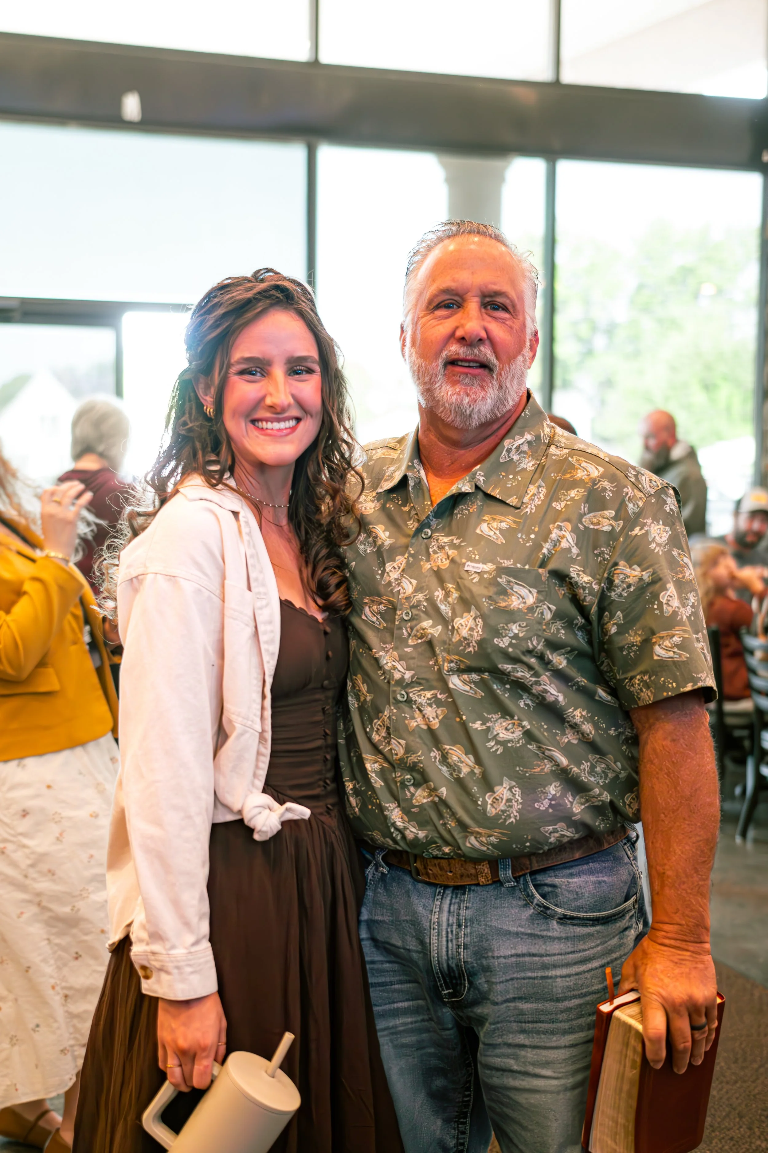 A smiling young woman with curly brown hair and a man with gray hair and beard are standing close together indoors. The woman is wearing a beige jacket over a brown dress, and the man is wearing a patterned short-sleeve shirt and holding a wooden book and a beverage cup with a straw. There are other people in the background.