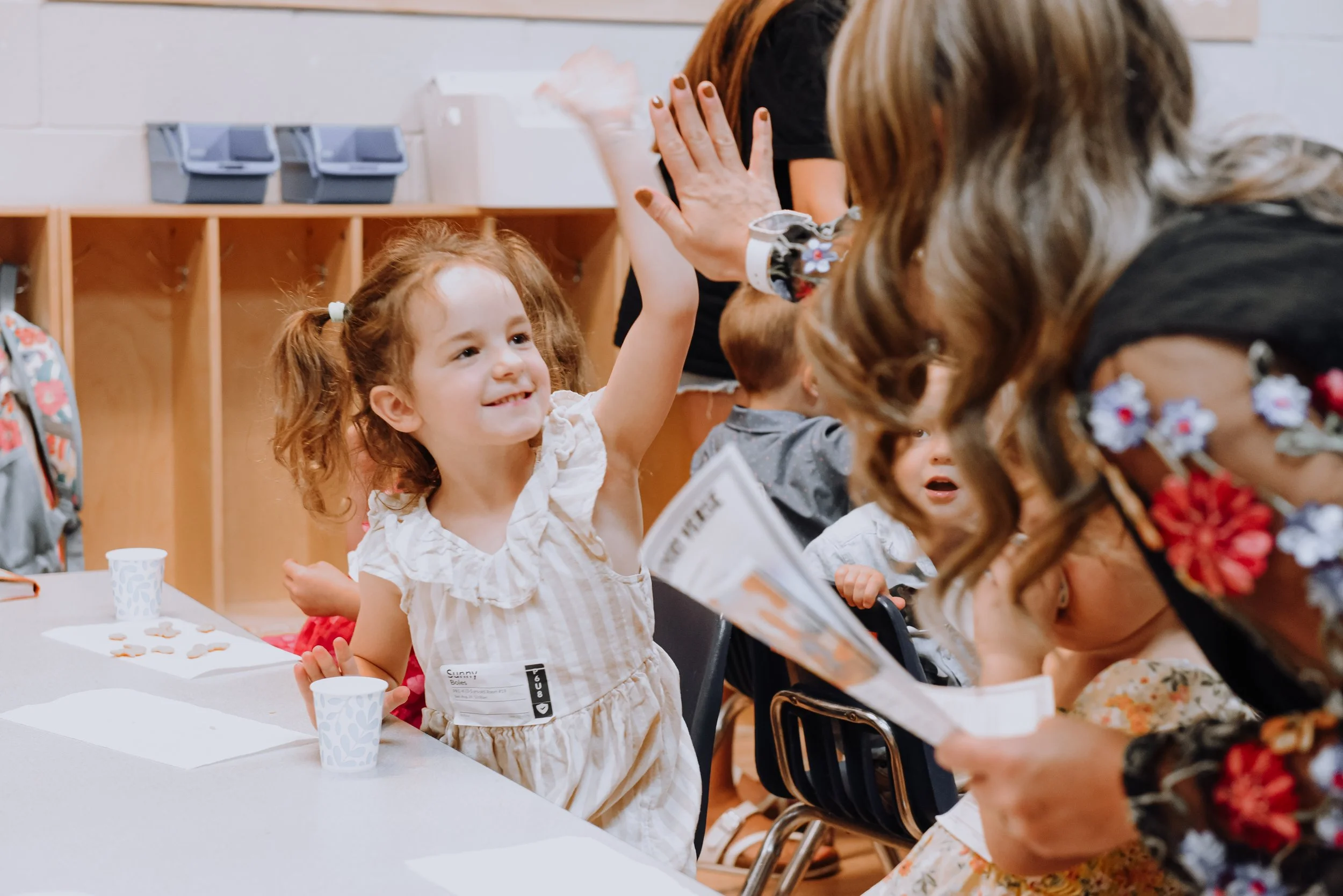 A young girl with curly red hair and a striped dress raises her hand during a classroom activity, smiling. Other children and adults are present, engaged in the event, with some adults leaning in and talking to the children.