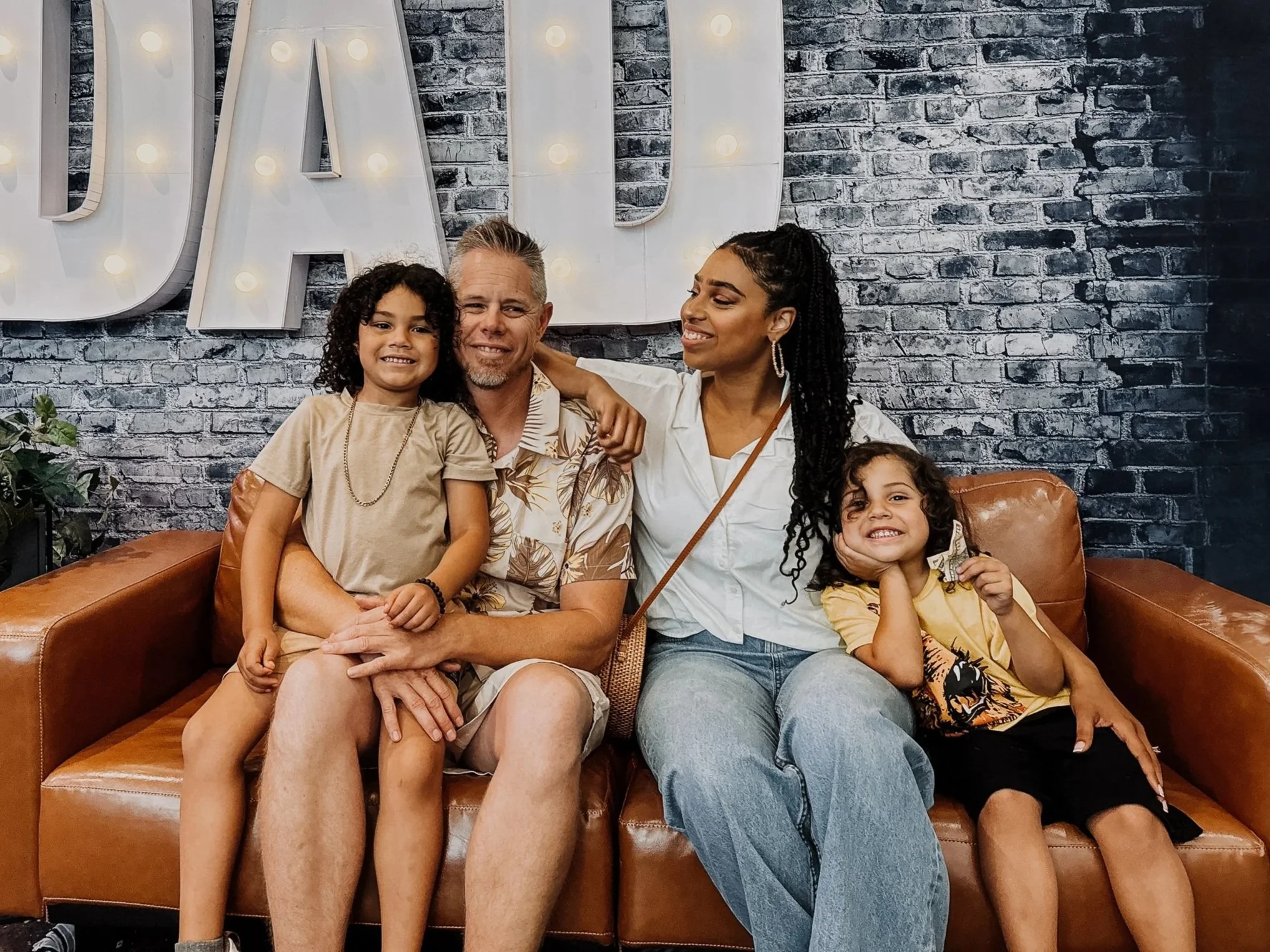 A group of four people, including two women and two young girls, sitting on a brown leather couch in front of a gray brick wall with large illuminated letters spelling 'LADY' behind them, smiling and close together.