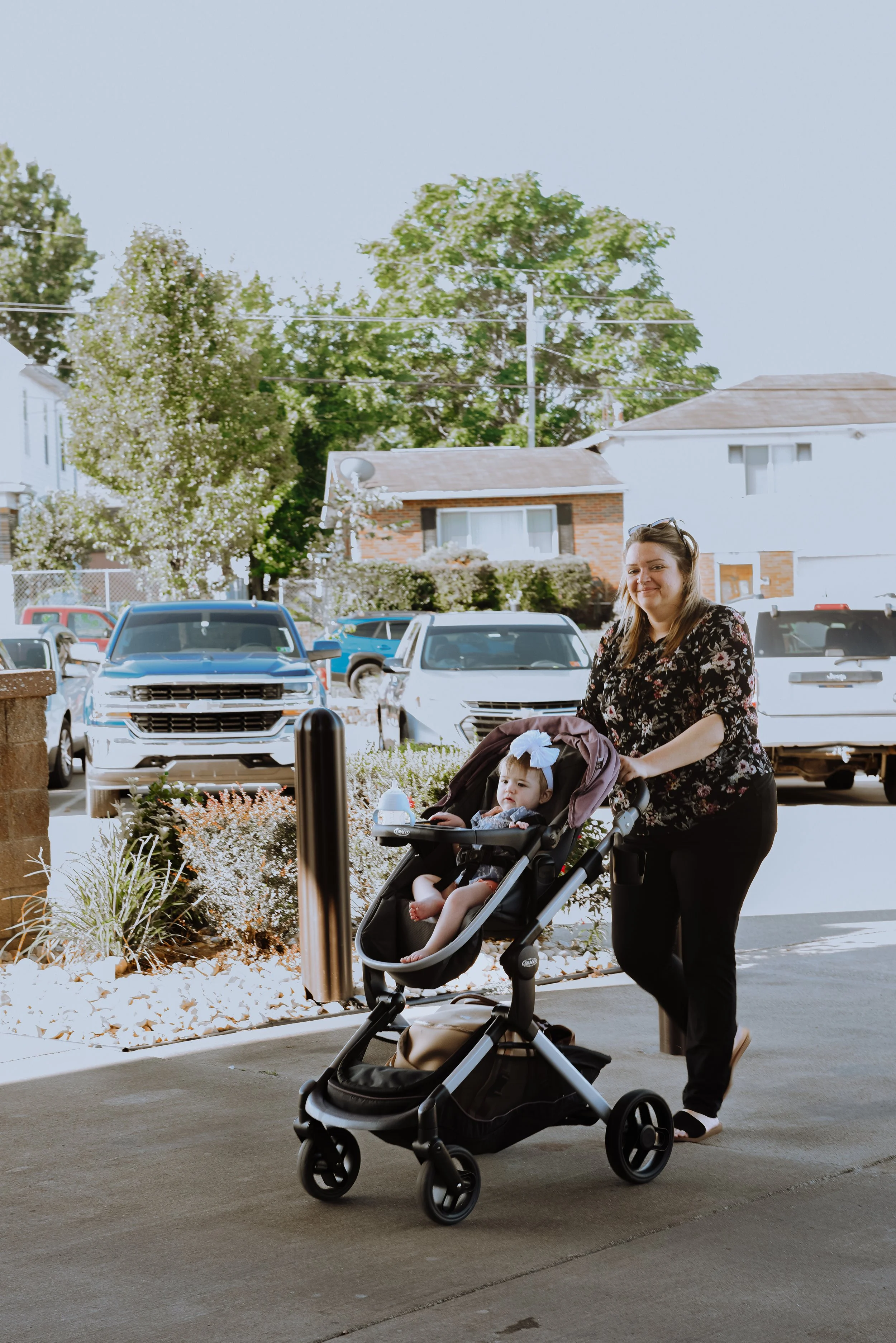 A woman walking with a stroller containing a young girl in a black dress with a white bow, on a sidewalk outside near parked cars and trees in the background.