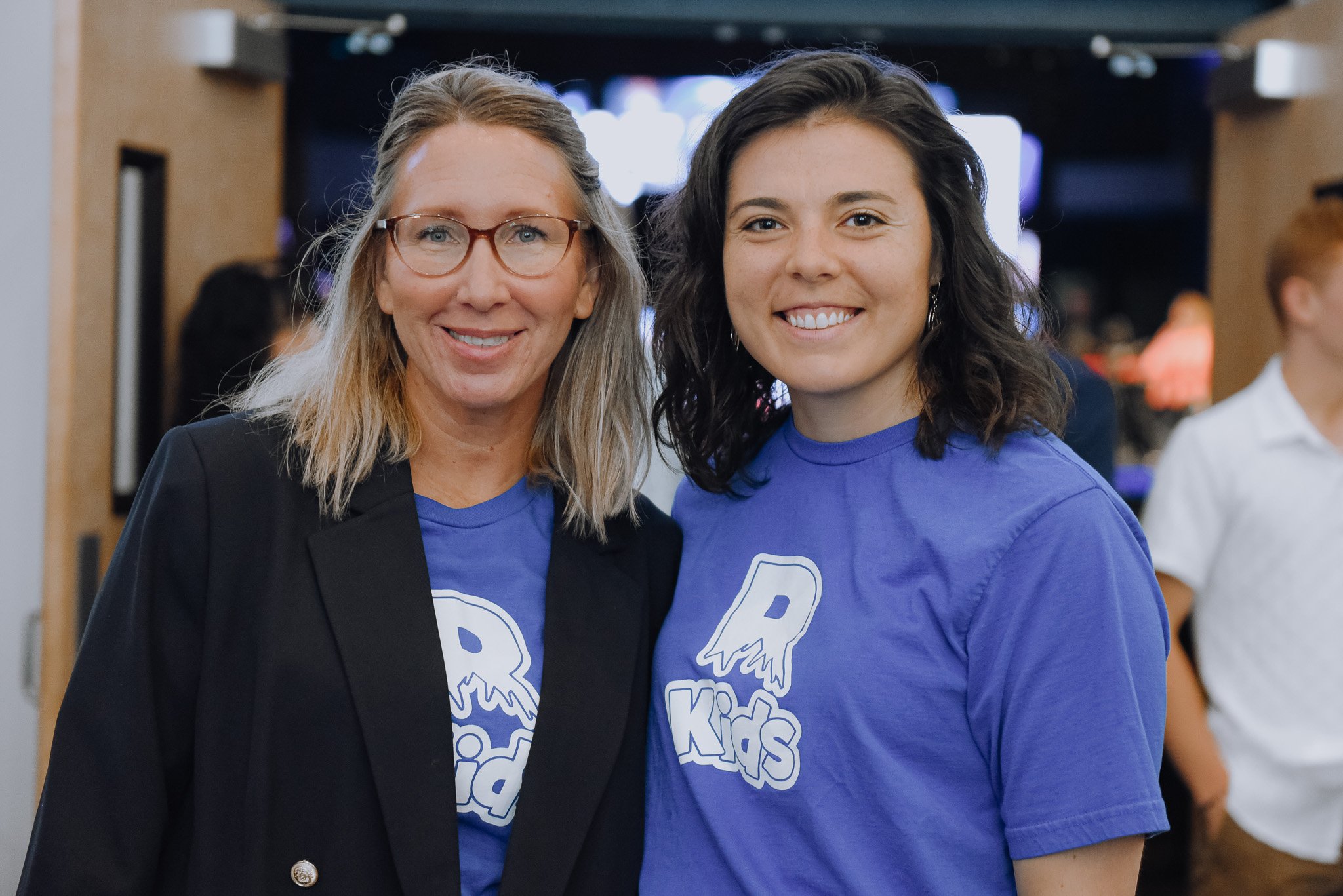 Two women smiling at the camera, one with glasses and long gray hair, wearing a black blazer, and the other with dark wavy hair, wearing a blue T-shirt with a logo, in an indoor event setting.