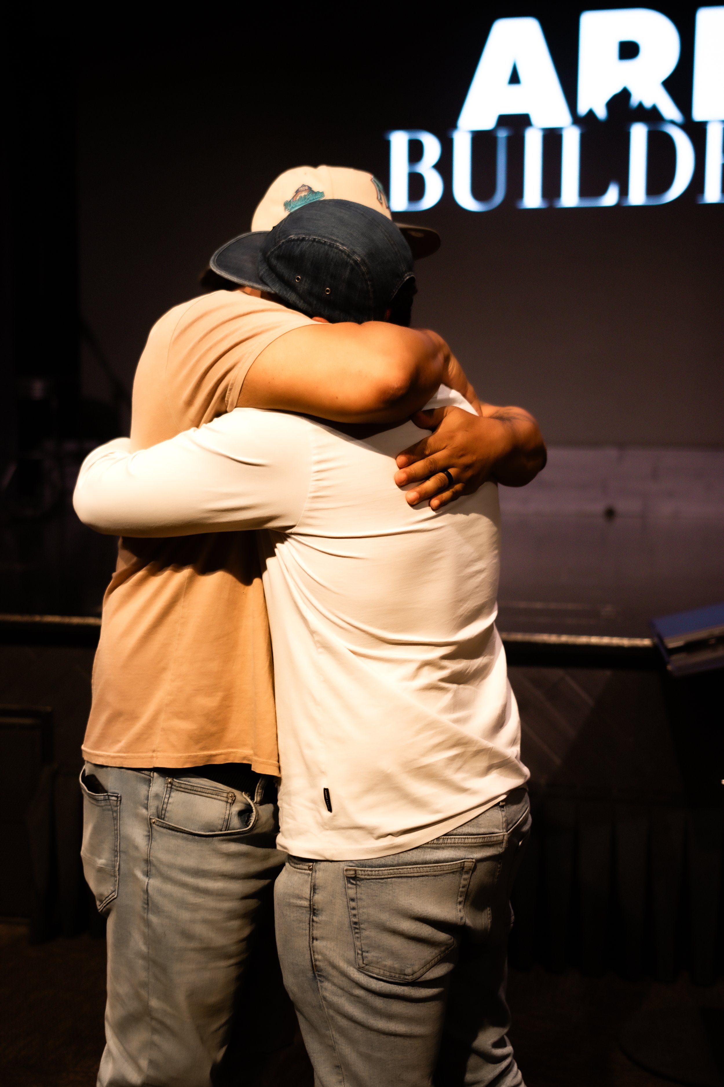 Two people hugging on stage with a dark background, and a partially visible sign that says 'ART' and 'BUILD'.