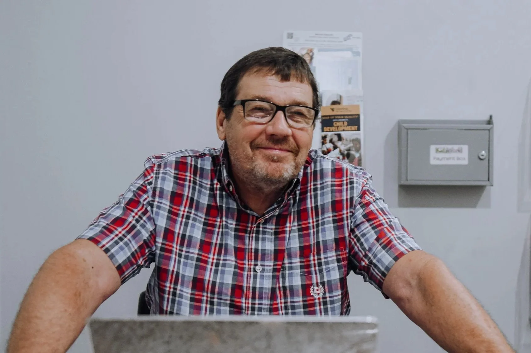 A middle-aged man with glasses and a beard, wearing a red plaid shirt, sitting at a desk and smiling at the camera in an office setting.