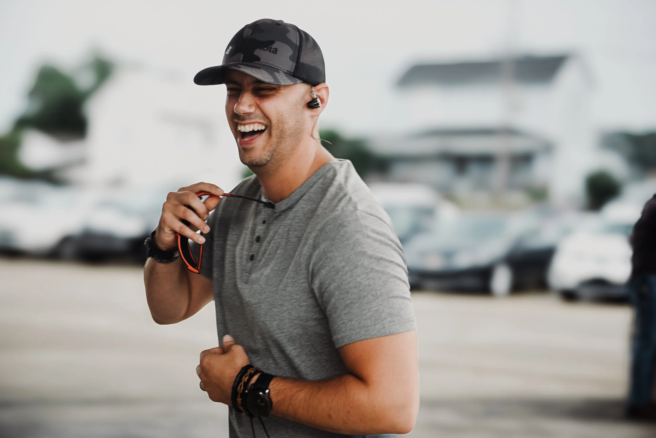 A man running outdoors wearing a gray t-shirt, a black cap, and headphones, smiling and holding sunglasses in his hand.