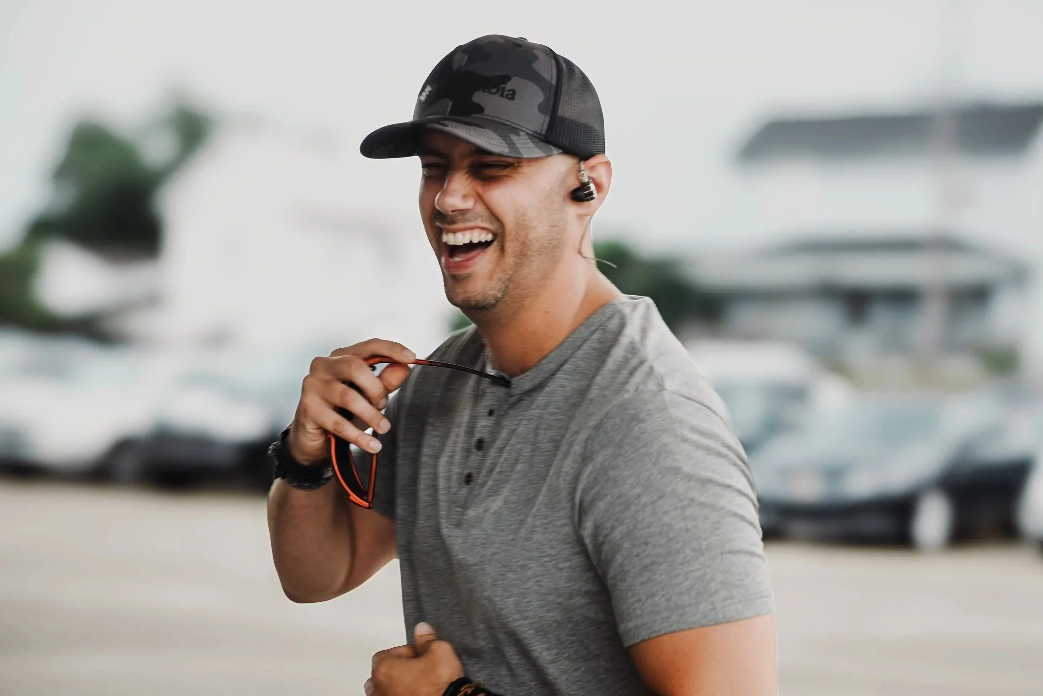 A young man wearing a camouflage baseball cap, a gray athletic shirt, and black wireless earbuds, smiling while running outdoors.