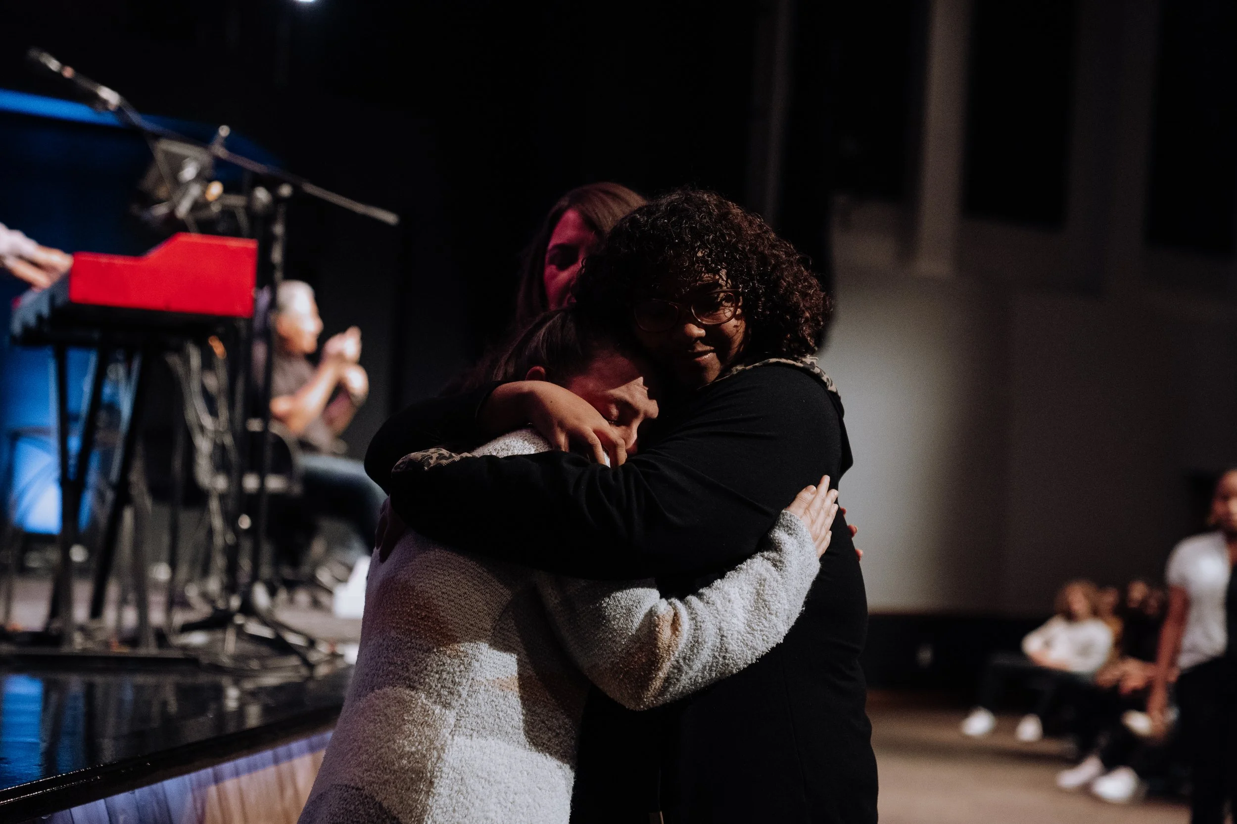 Two women sharing a hug on stage in a theater, with an audience in the background.