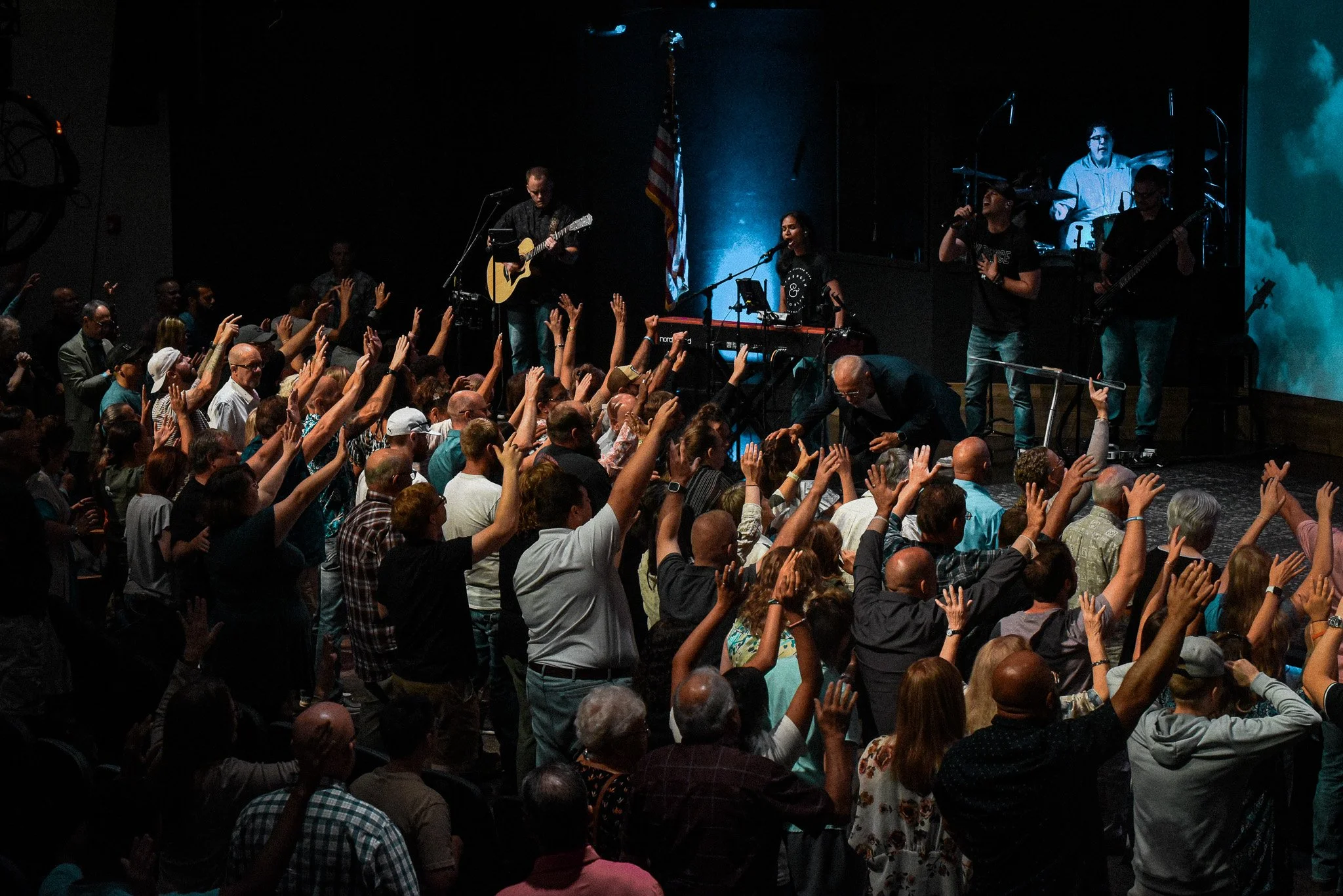 A large crowd of people raising their hands while a band performs on stage in a dark venue. The band has multiple members, with one playing guitar, another singing into a microphone, and others playing instruments. An American flag is visible near the stage.