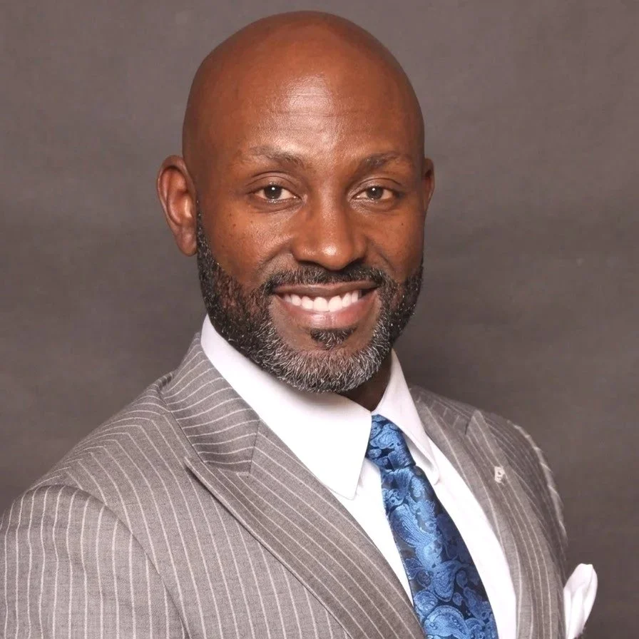 Headshot of a smiling Black man wearing a gray pinstripe suit, white shirt, and blue patterned tie.
