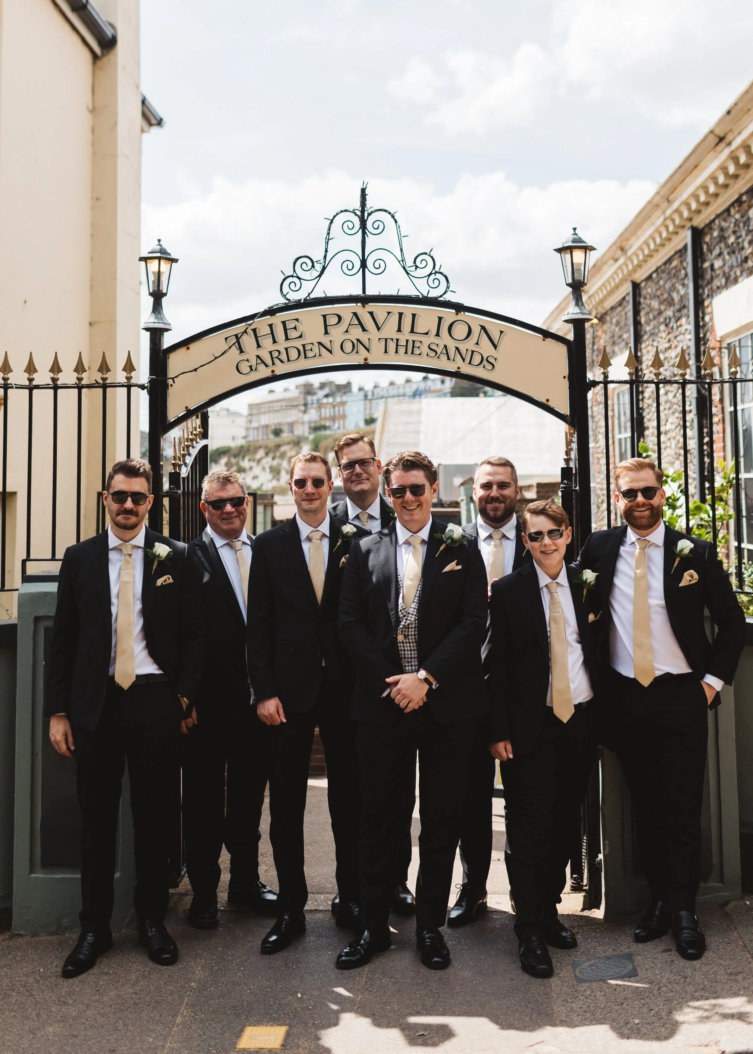Group of men dressed in formal suits and ties, standing under an archway with a sign that reads "The Pavilion Garden on the Sands", at an outdoor event.