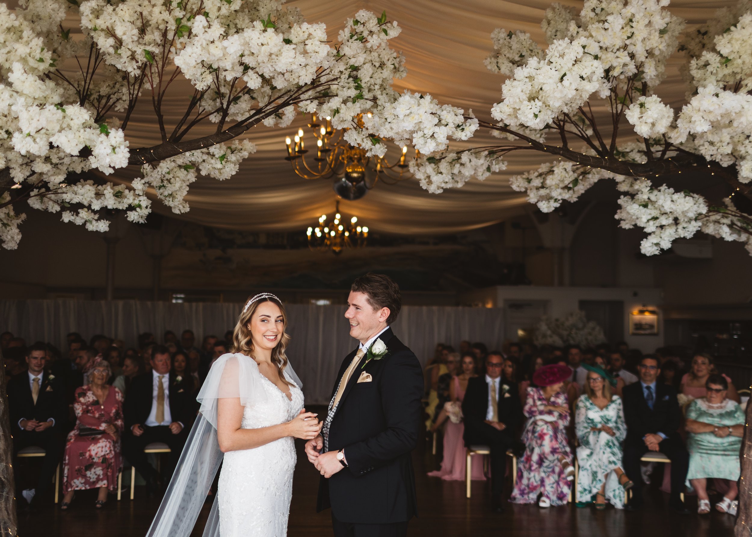 A bride and groom exchanging vows under a ceiling decorated with white flowers and chandeliers during a wedding ceremony, with seated guests in the background.