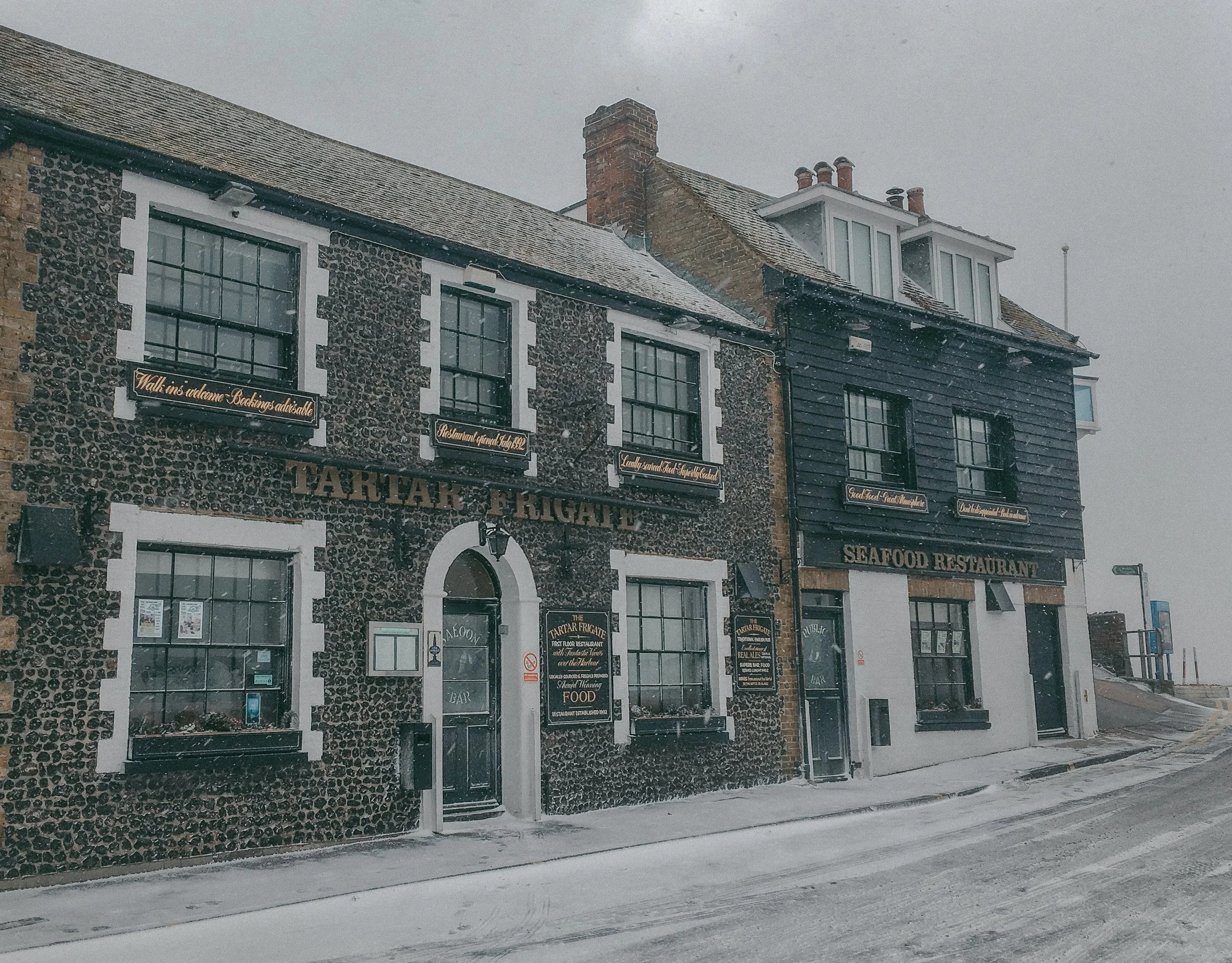 A row of brick buildings with large black-framed windows during snowy weather, with signs indicating a seafood restaurant and other shops on the ground floor.
