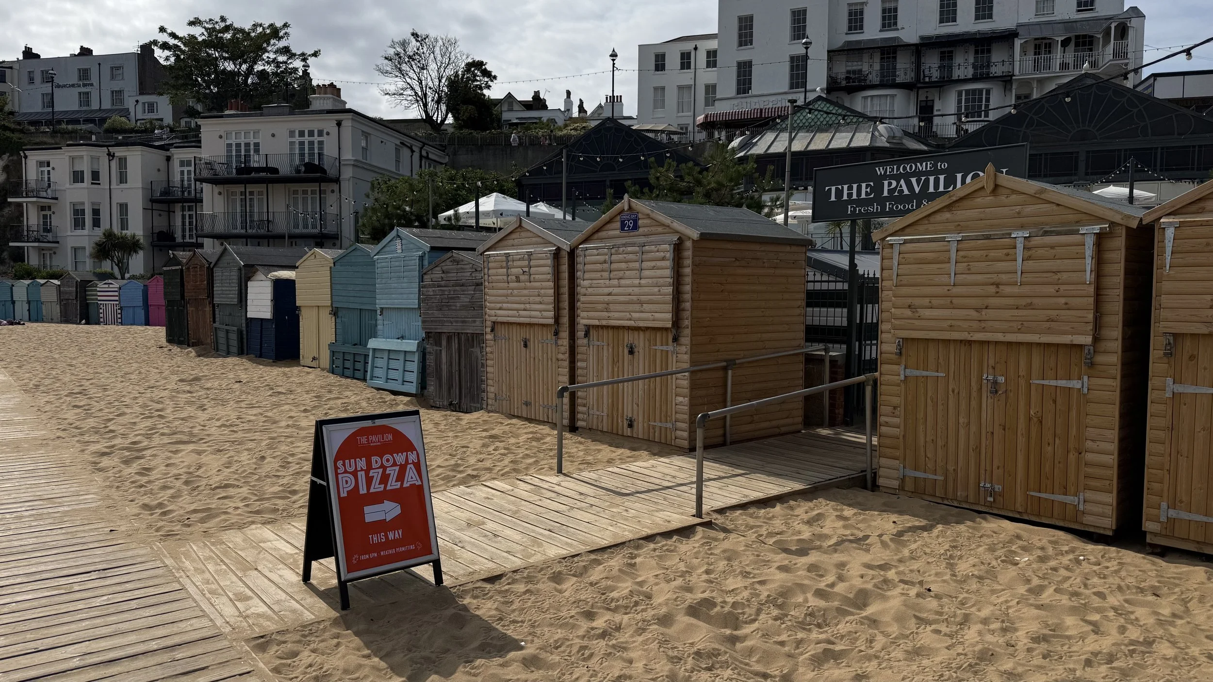 View of colorful beach huts on sand with a boardwalk and wooden ramp, sign advertising pizza, and buildings in the background, cloudy sky.