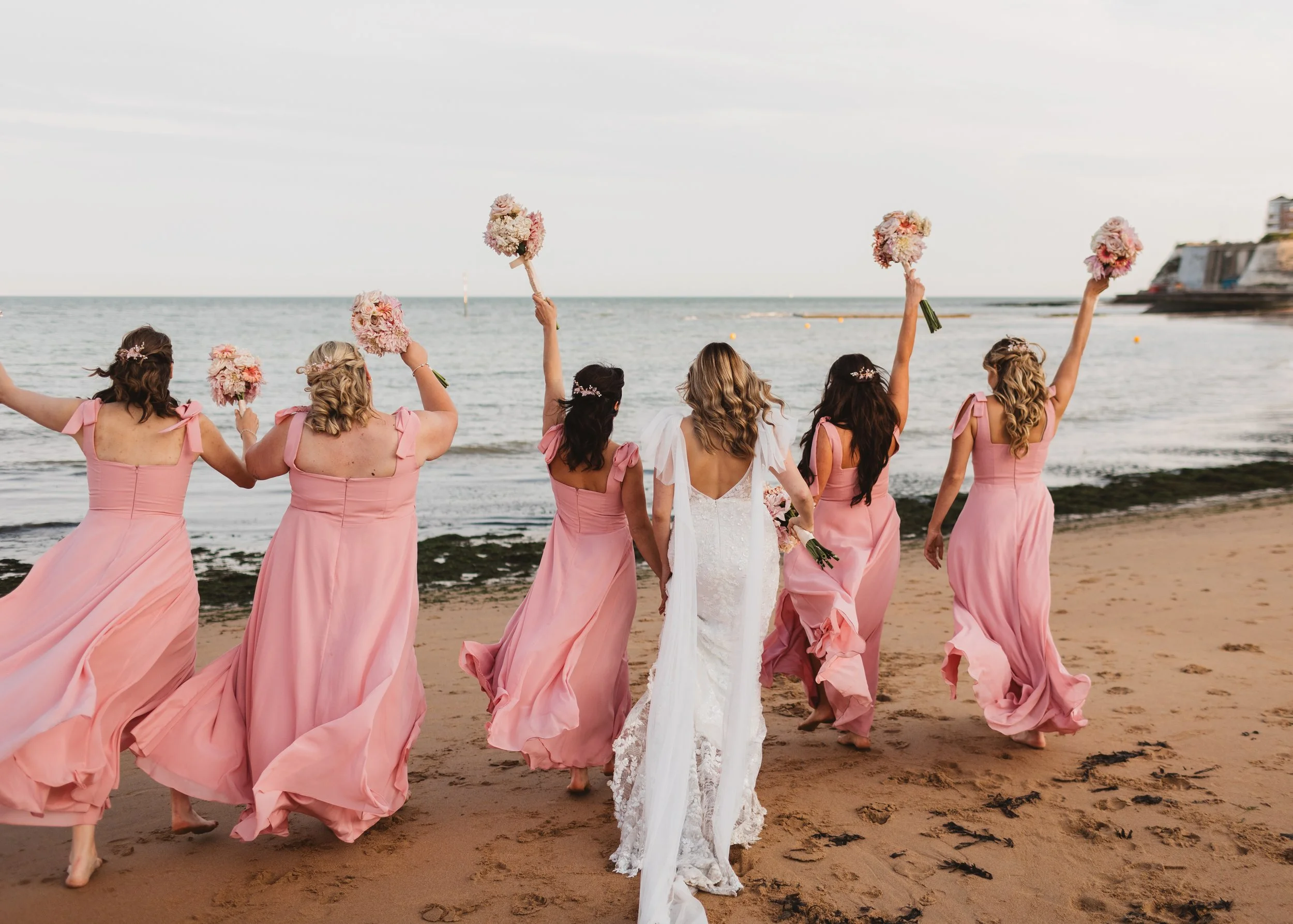 A group of women wearing pink dresses and holding bouquets walk along the beach near the ocean, with some of them raising their bouquets in the air. A bride in a white wedding dress is among them, walking barefoot on the sand.