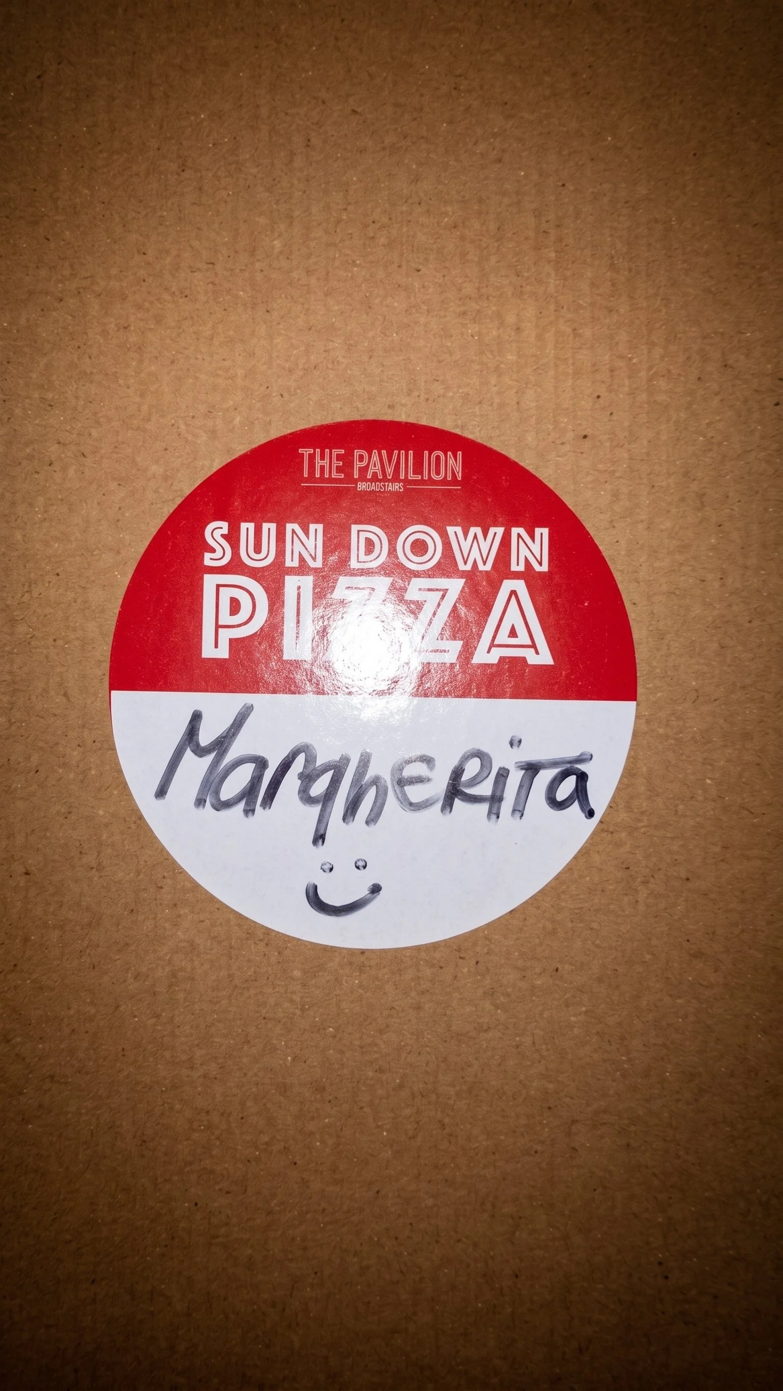 A pizza box with a round sticker on it. The sticker says 'Sun Down Pizza' at the top in red text, and 'Margherita' handwritten in black marker with a smiley face below.