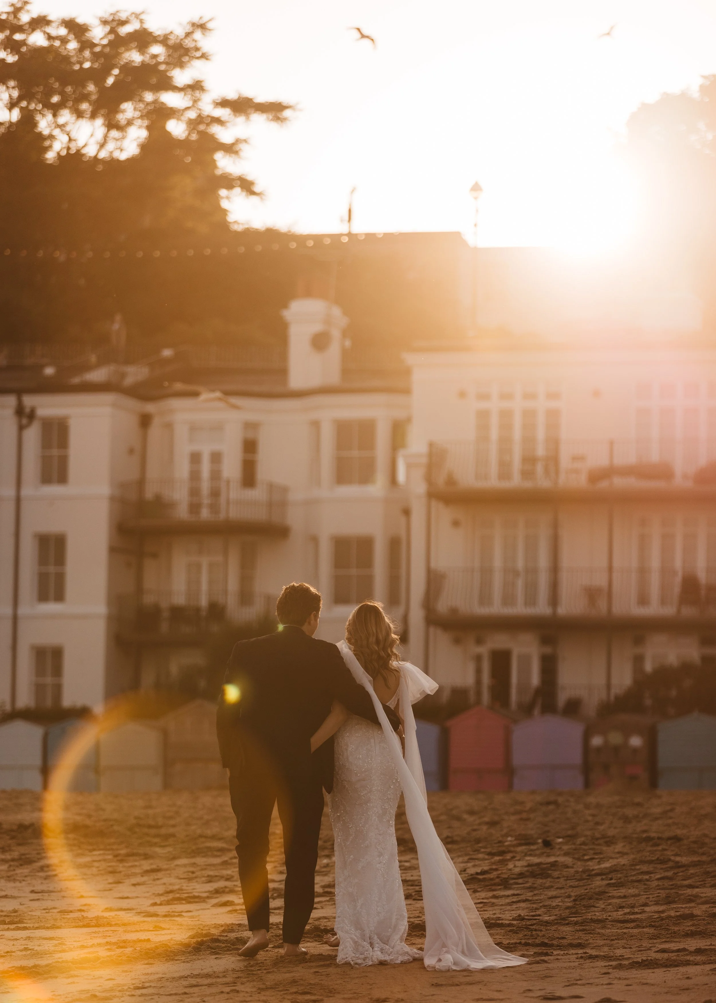 A couple dressed in wedding attire walking on a beach at sunset, with the woman in a white wedding gown and the man in a tuxedo, and a large white building with balconies in the background.