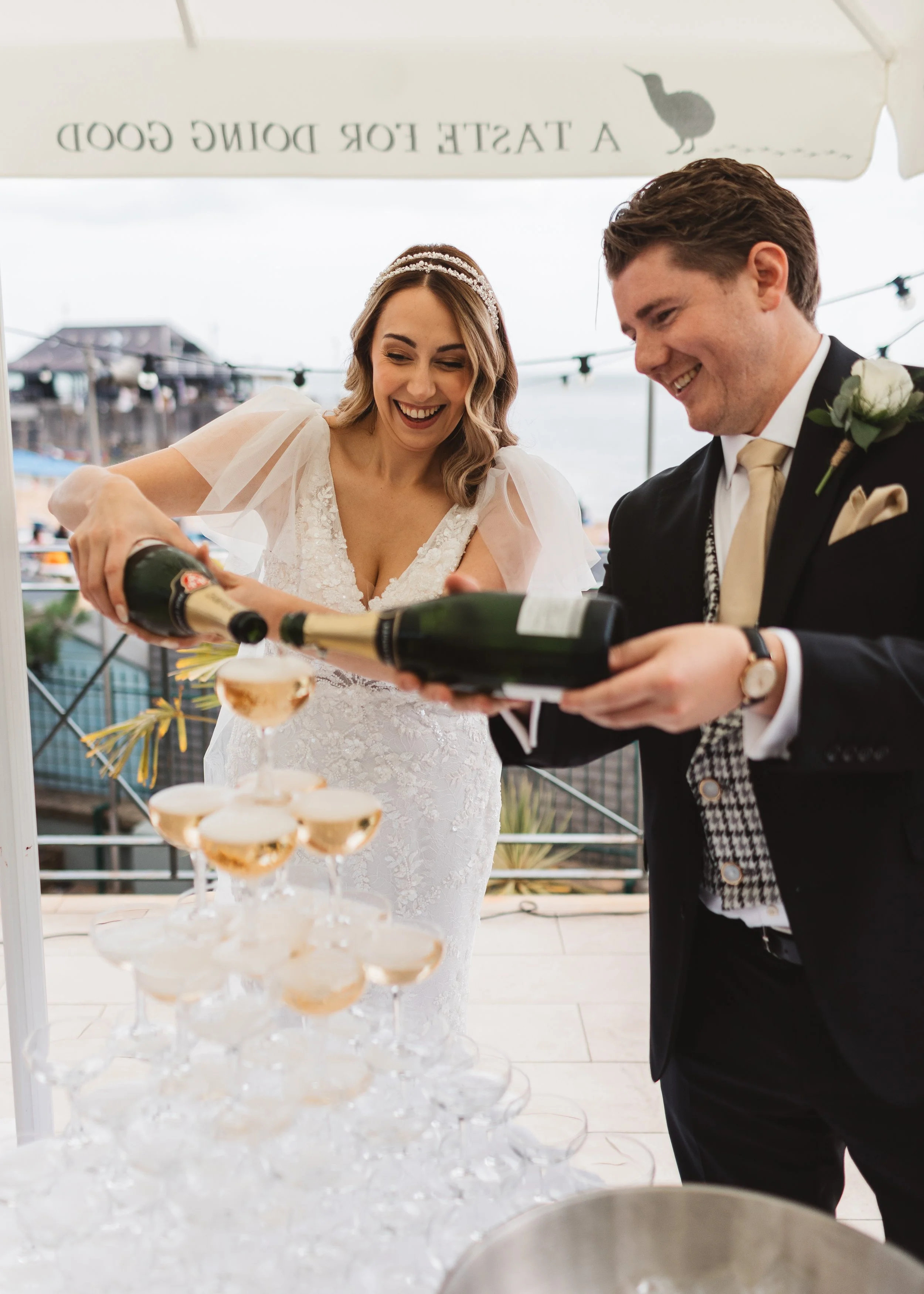 A bride and groom at a wedding reception, pouring champagne into a tower of glasses on a balcony with a view of a pier and water in the background.