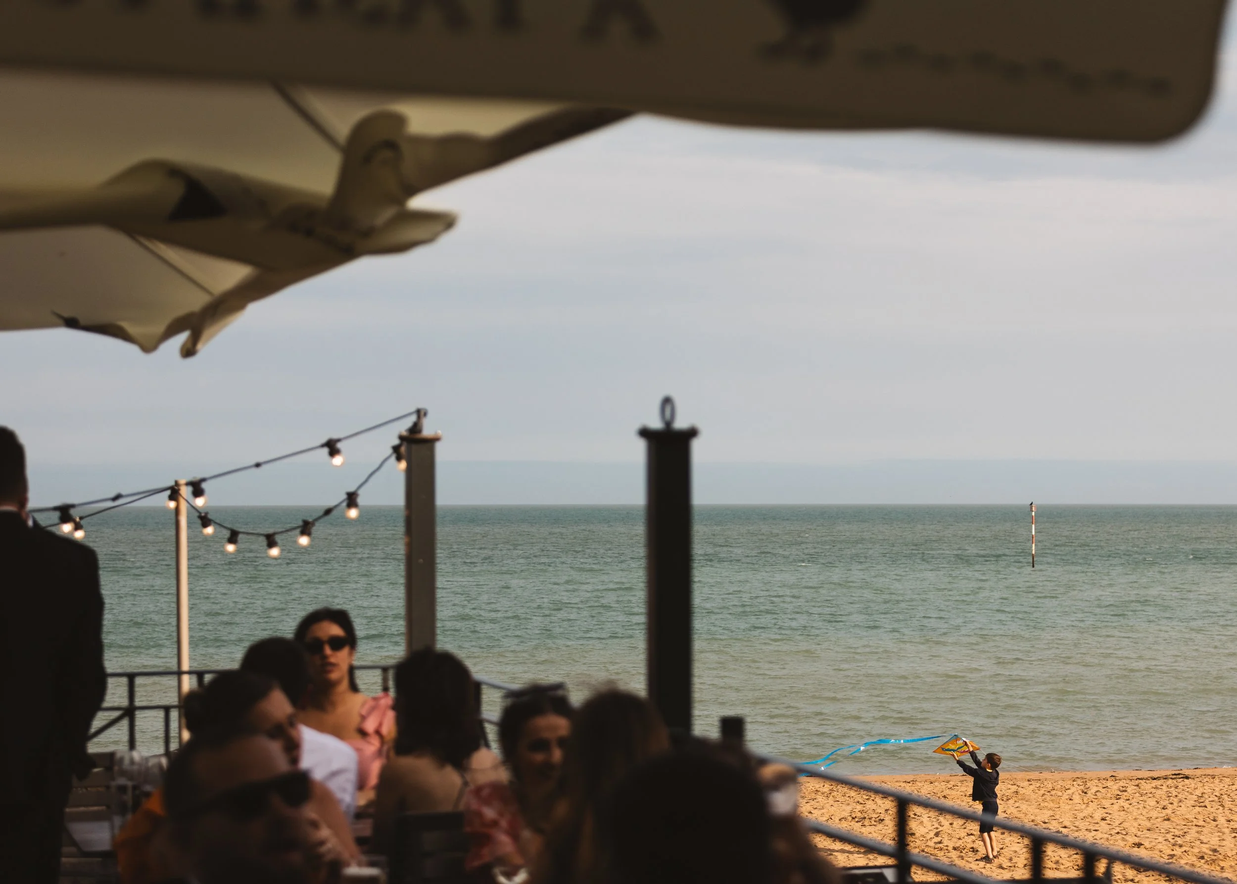 People sitting on a beachside patio, some wearing sunglasses, with a view of the ocean in the background. There is a child flying a kite on the beach, and an umbrella partially visible in the foreground, along with string lights hanging above.