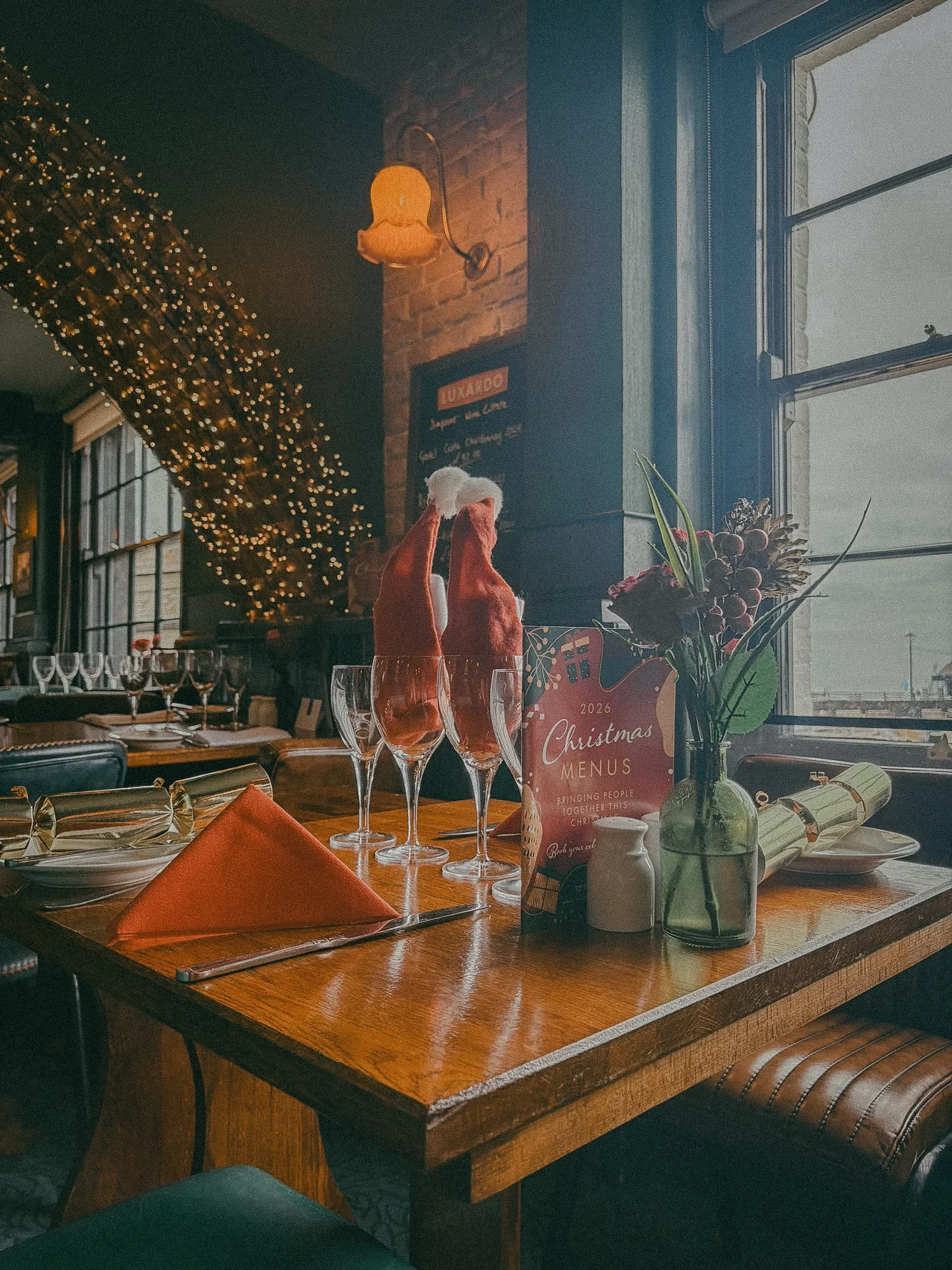 A decorated restaurant table for Christmas with wine glasses, a festive menu, a small vase with flowers, and holiday decorations, near a window with natural light.