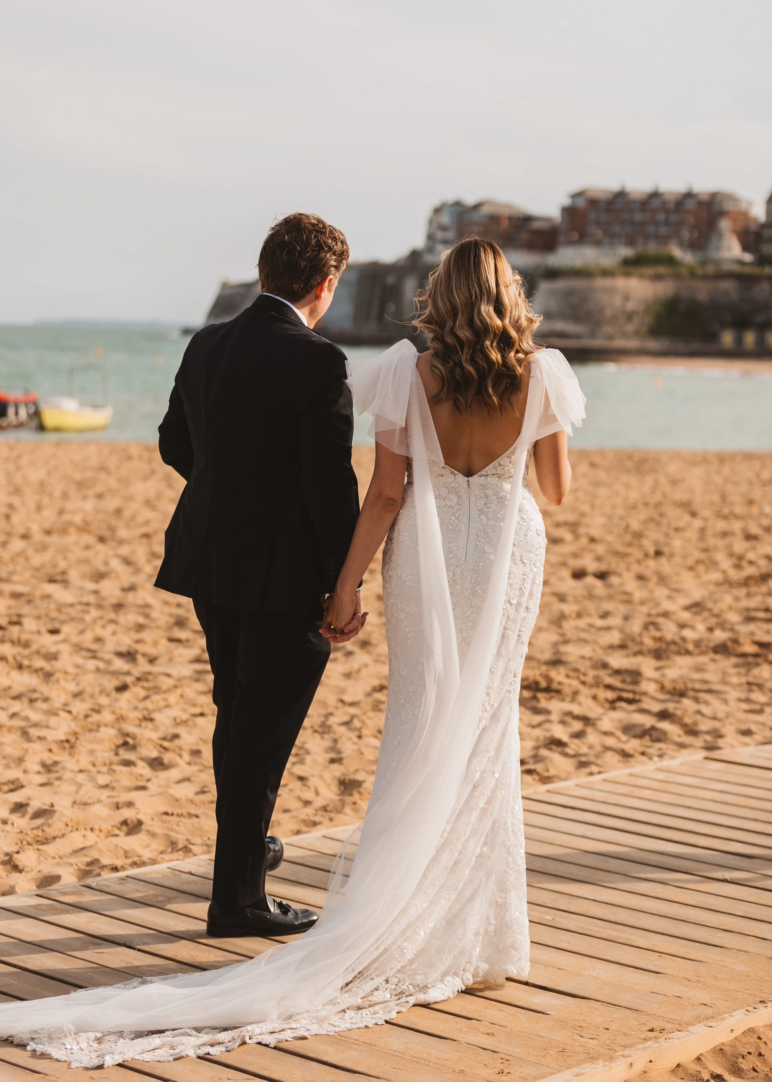 A bride and groom holding hands on a beach, facing away, with a city and ocean in the background, during sunset.