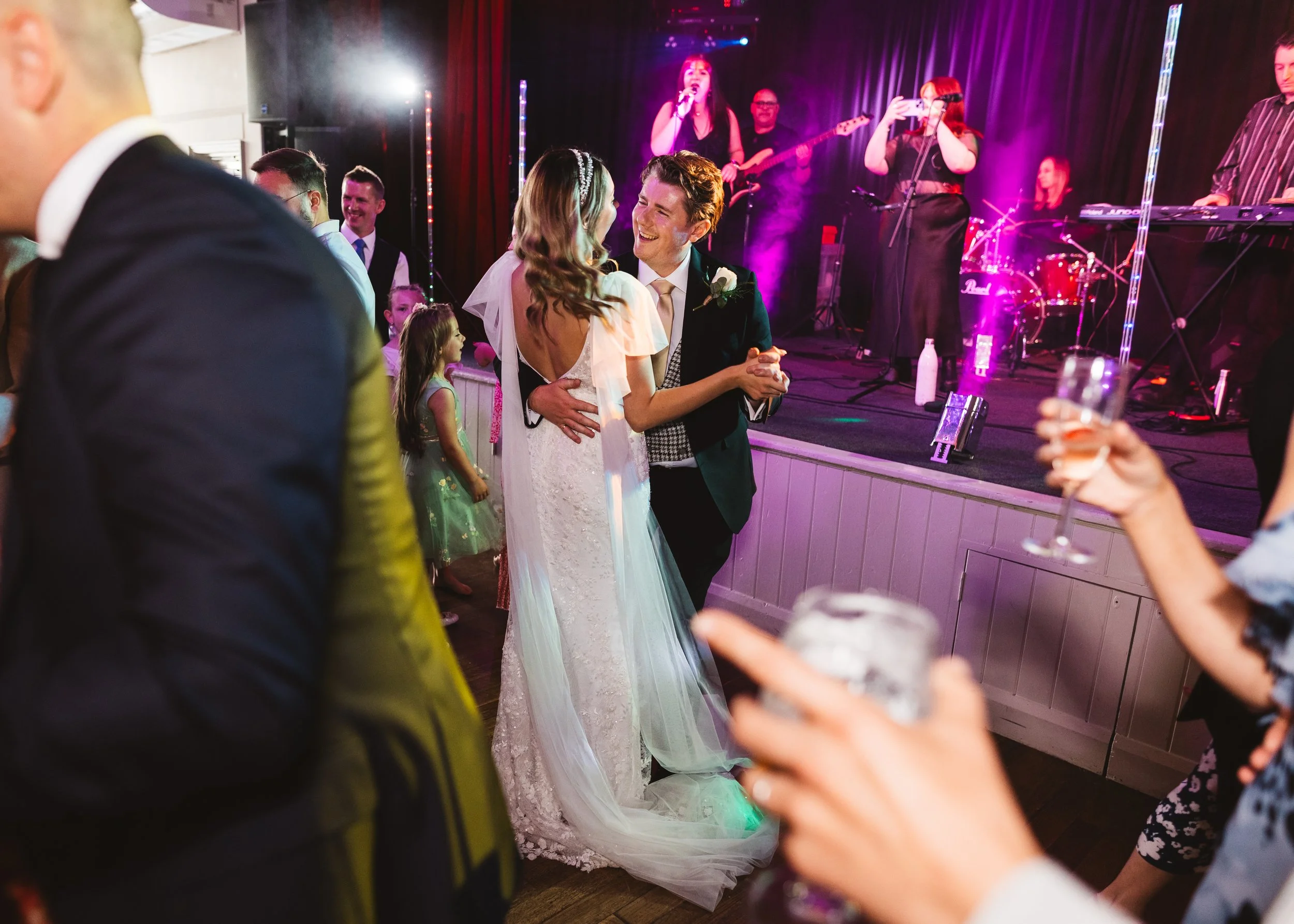 Bride and groom dancing during wedding reception, surrounded by guests, with band performing on stage in the background, purple and pink lighting.
