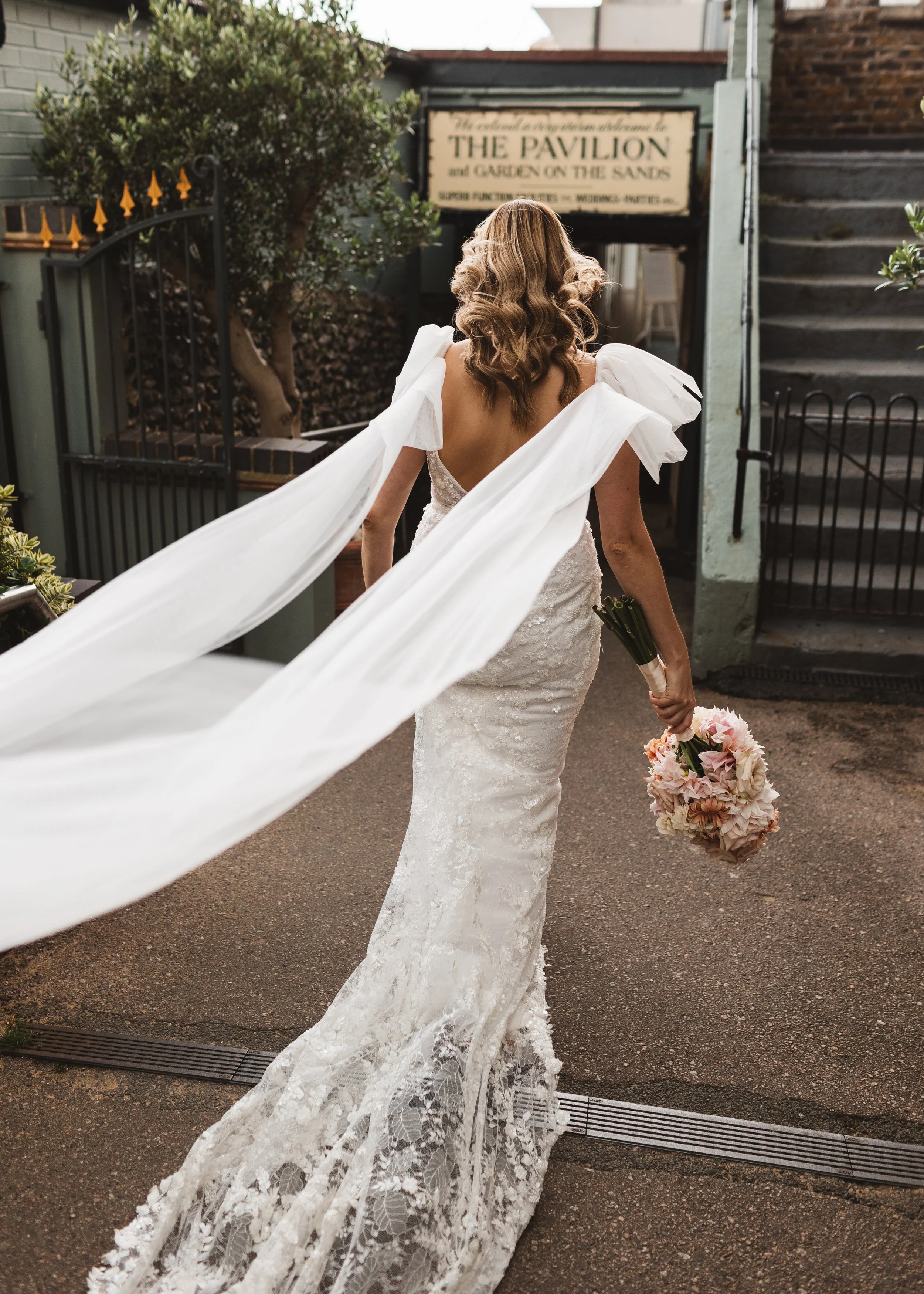 A bride wearing a white lace wedding gown with puffed sleeves walking away from the camera, holding a bouquet of pink and cream flowers, with a sign and stairs in the background.