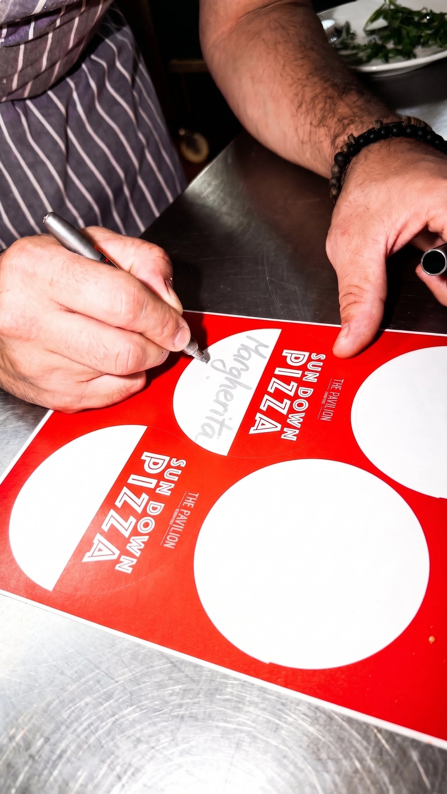 A person signing a pizza box label with a silver marker. The box is red and reads 'Sun D'W' and 'The Pavilion'.