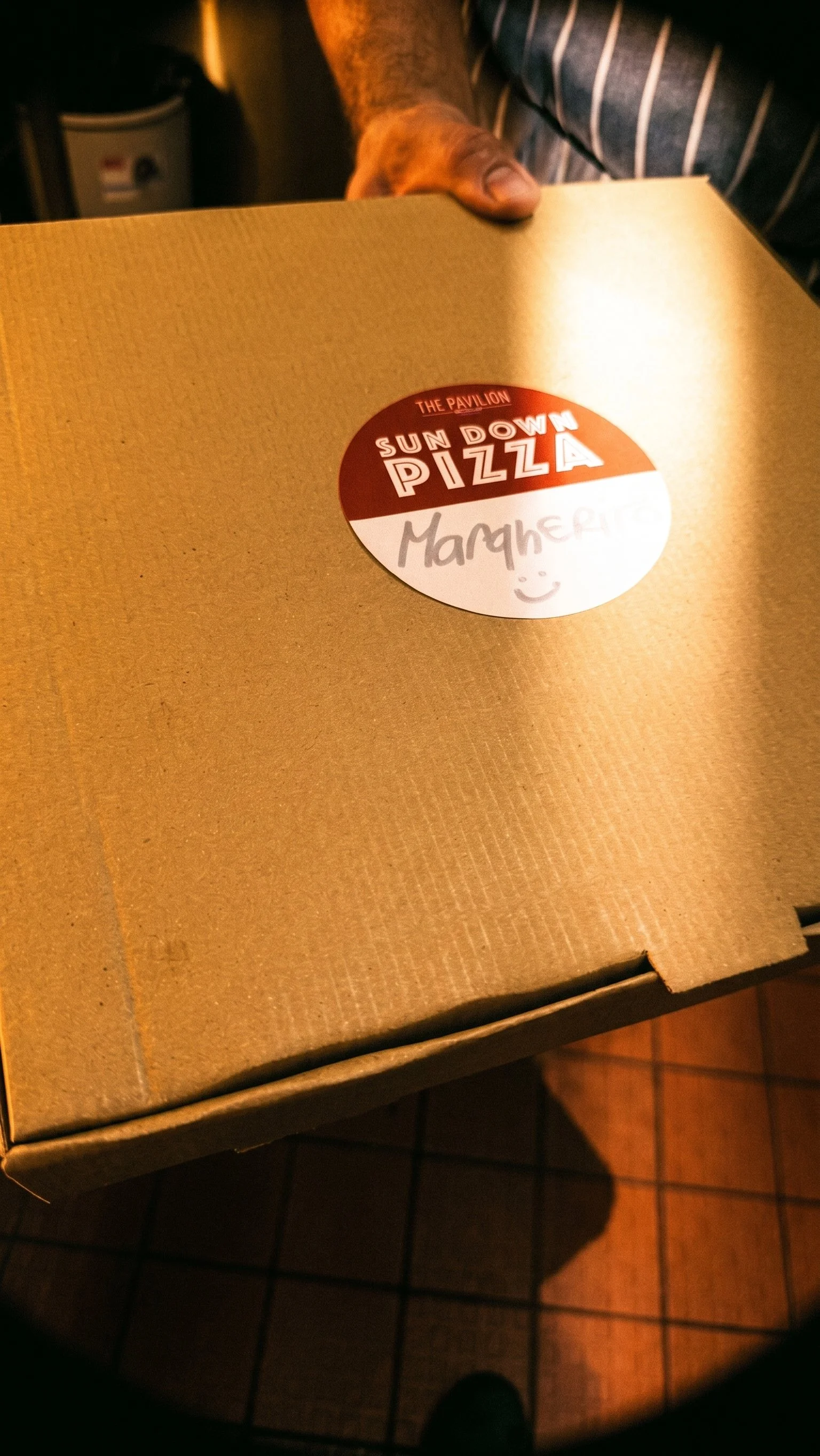 A closed cardboard pizza box labeled 'Sun Down Pizza' with a handwritten note saying 'Margerita' and a smiley face on a sticker.