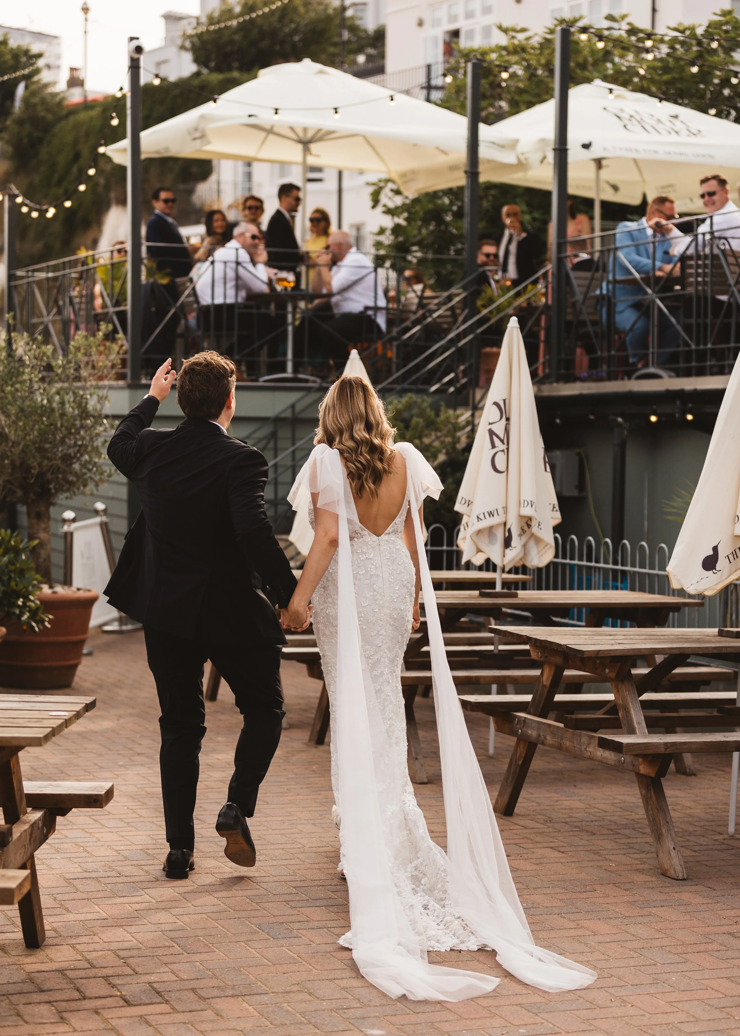 A couple in wedding attire holding hands as they walk away from a gathering on a patio, with guests sitting at tables under umbrellas in the background.
