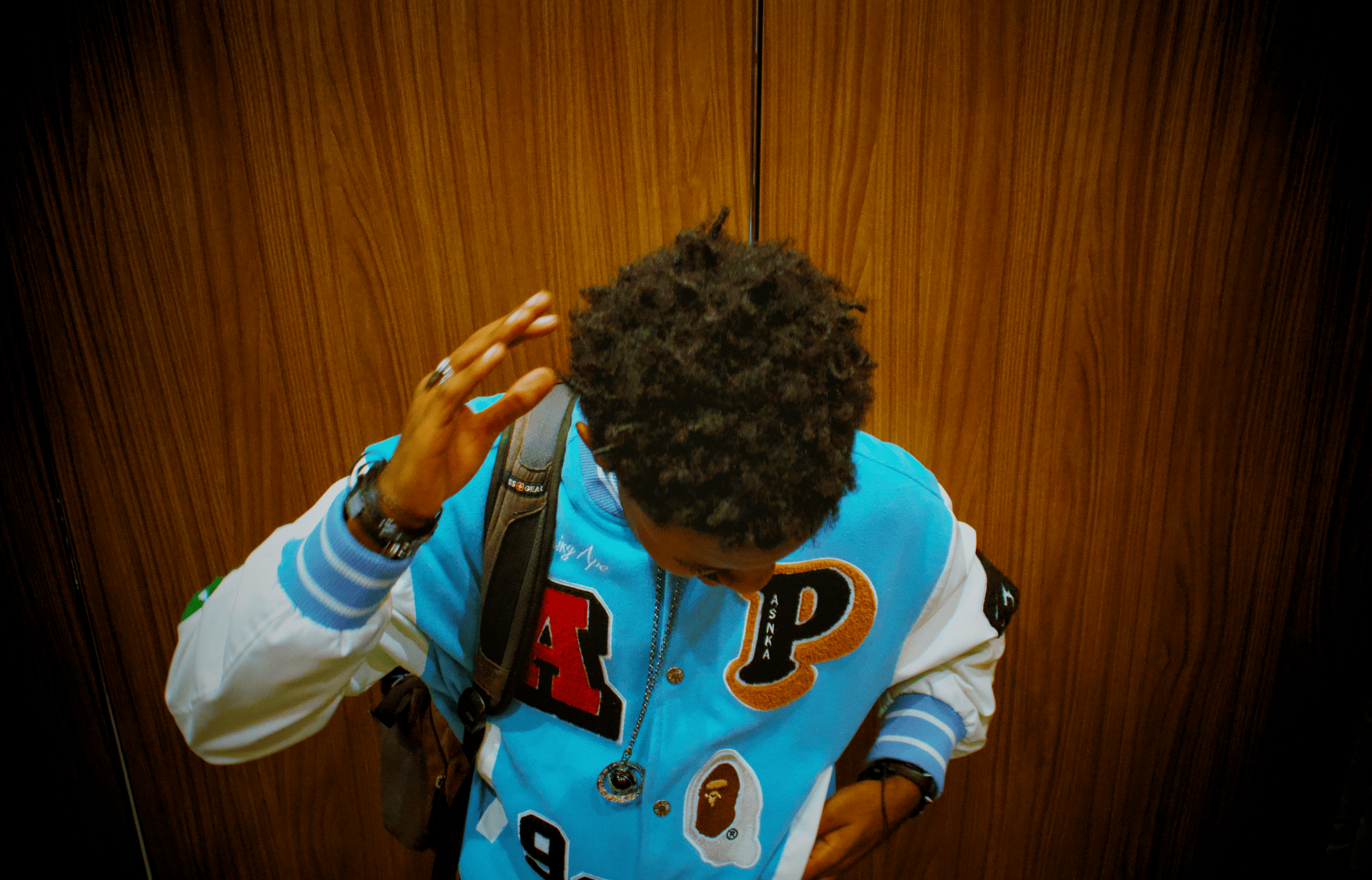 Person with curly hair wearing a colorful varsity jacket and a backpack, standing in an elevator with wood-paneled walls.