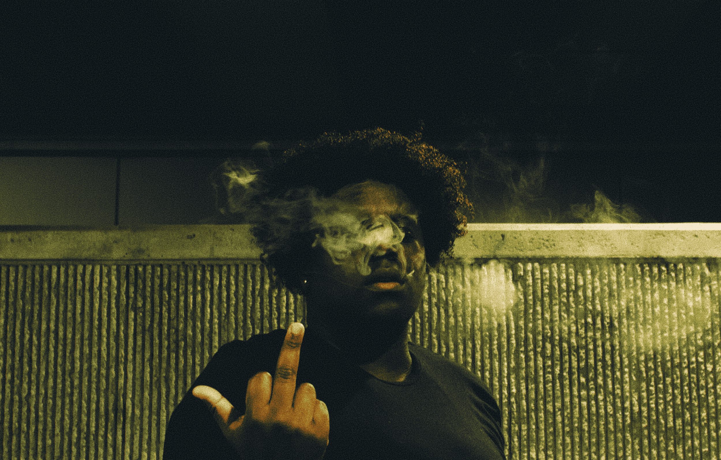 A person with curly hair and dark clothing is standing behind a metal fence at night, smoking and holding up their middle finger.
