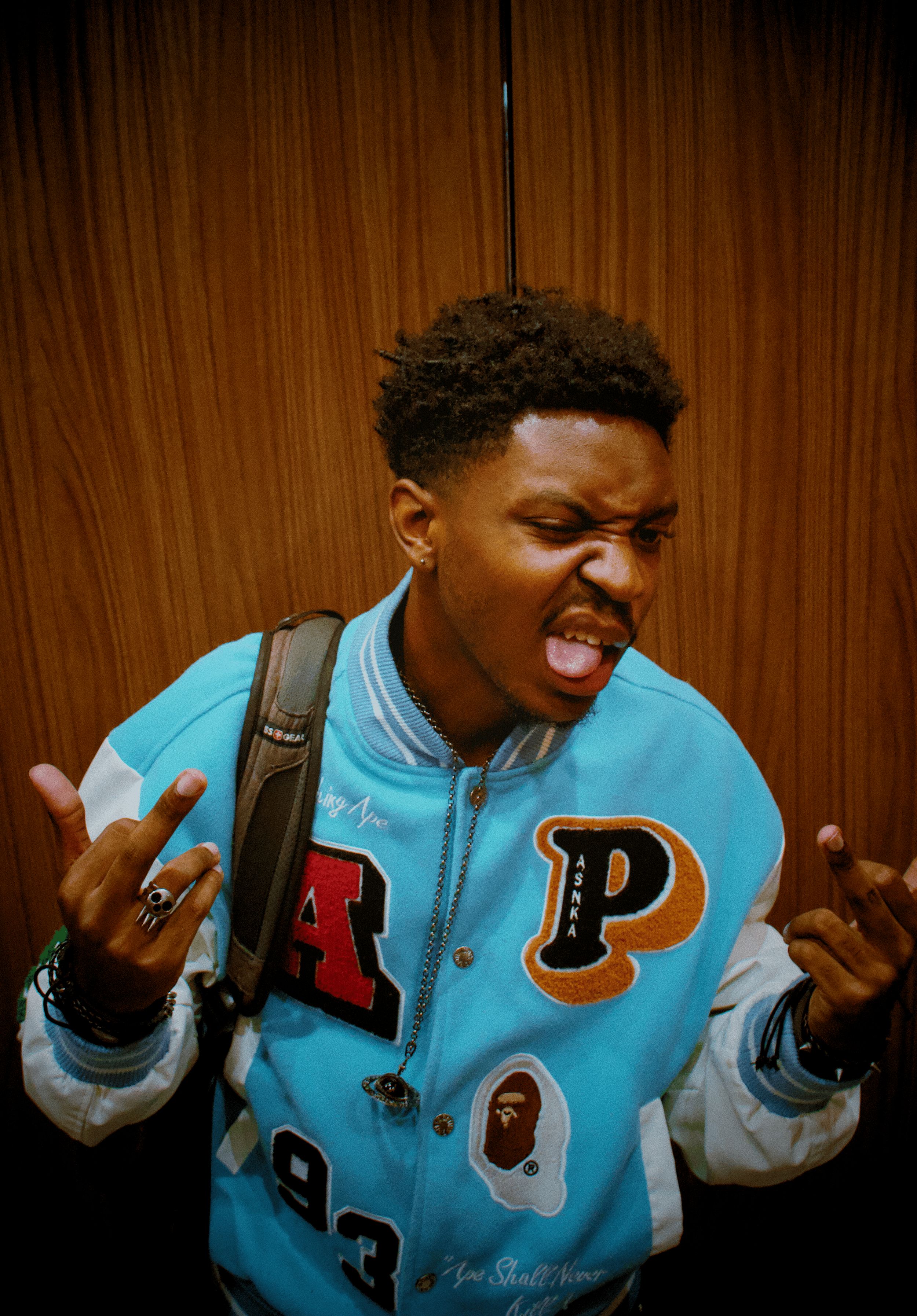 A young man with brown skin and curly hair making a playful face and pose, with one eye closed and tongue out, in front of a wooden wall. He wears a blue jacket with patches and letters, and has various accessories including rings, bracelets, and a n