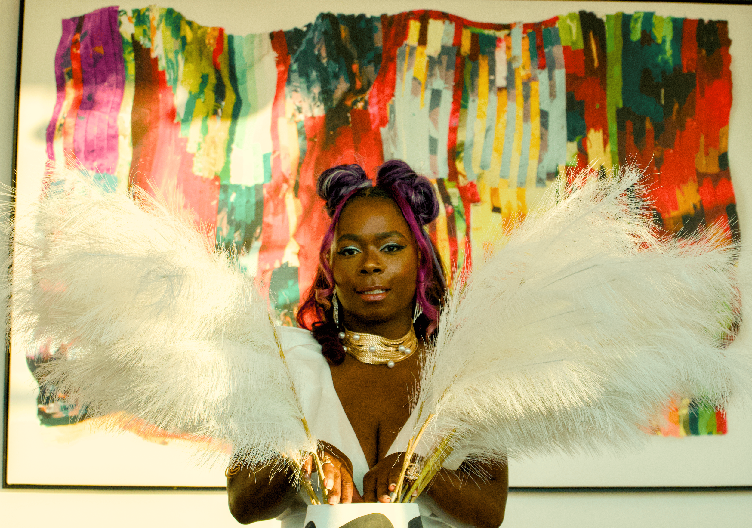 A woman with purple hair styled in two buns, wearing a pearl necklace and an off-the-shoulder white outfit, holding large white feather fans. Behind her is an abstract colorful painting.