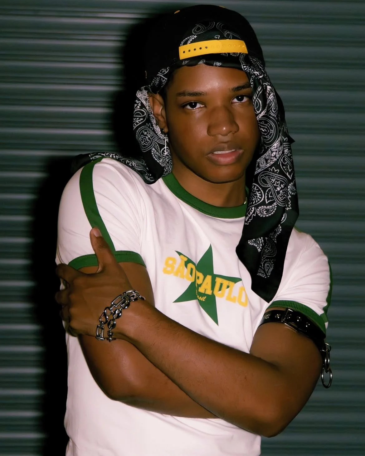 Young man wearing a white T-shirt with green accents, a black and yellow backward cap, and a black bandana, standing with arms crossed in front of a blurred background.