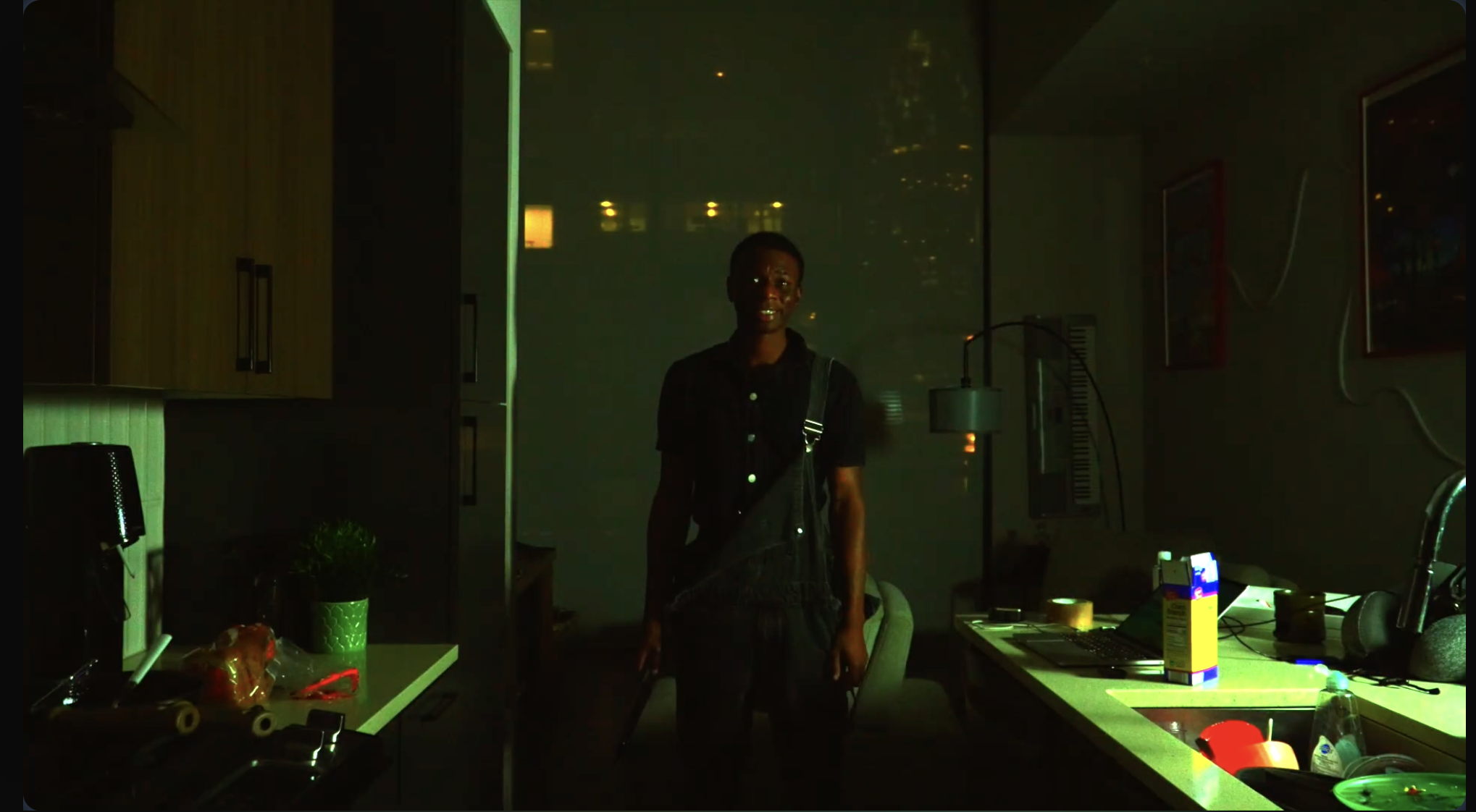 A woman standing in a dimly lit room with a greenish glow, possibly from a laser or light source, with various objects on a counter including a box of tissues, a bowl, and a robot toy.