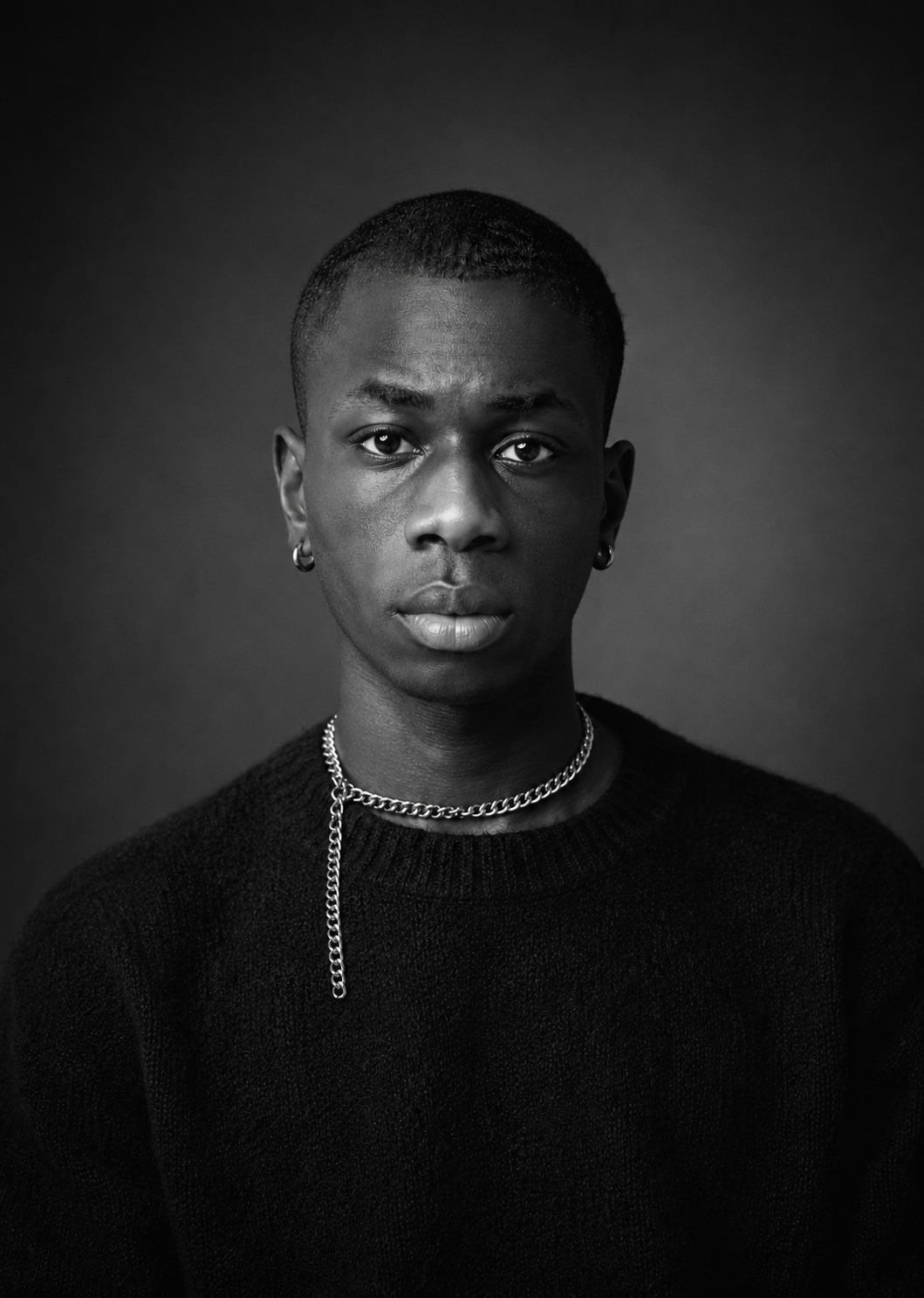 Black and white portrait of a young person with short hair, wearing earrings, a chain necklace with a dangling chain, and a dark sweater, looking directly at the camera.
