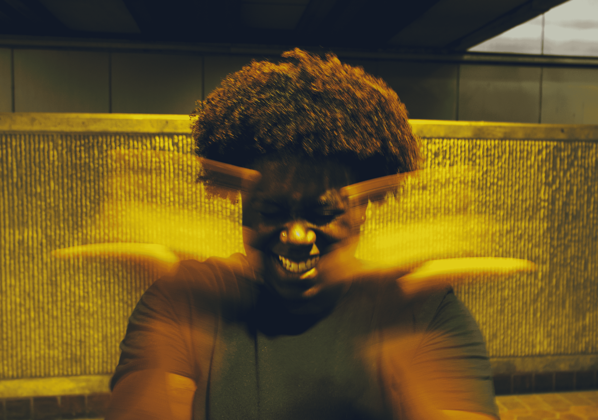 A person with dark skin and curly hair is smiling with teeth visible. They are wearing glasses and a dark top. The background is yellow with a textured pattern, and there's a structure above and behind them.