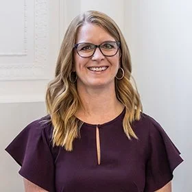 A smiling woman with shoulder-length blonde hair, wearing glasses and a dark purple top, standing in front of a neutral background.