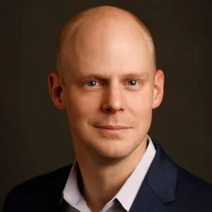 A professional headshot of a bald man wearing a dark suit and white shirt, against a dark background.