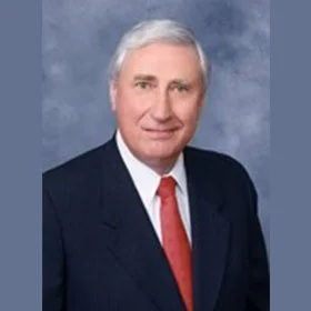 Portrait of an older man with gray hair, wearing a dark suit, white shirt, and red tie, against a gray background.