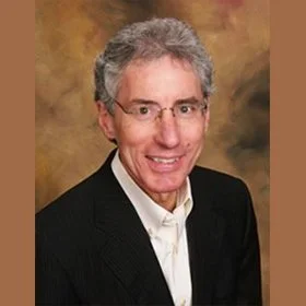 Portrait of an older man with gray hair and glasses, wearing a dark suit jacket and white shirt, smiling against a brown and gold background.