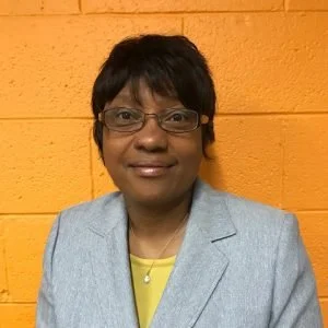 A woman with short black hair and glasses, wearing a light gray blazer over a yellow top, standing in front of a yellow brick wall.