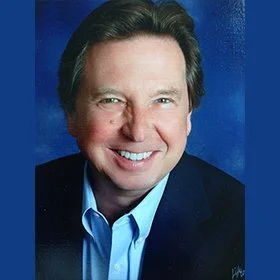 Close-up portrait of a smiling middle-aged man with dark hair, wearing a blue collared shirt and dark blazer, against a blue background.