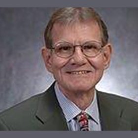 Portrait of an elderly man with glasses, wearing a gray suit, blue shirt, and patterned tie, smiling against a gray background.
