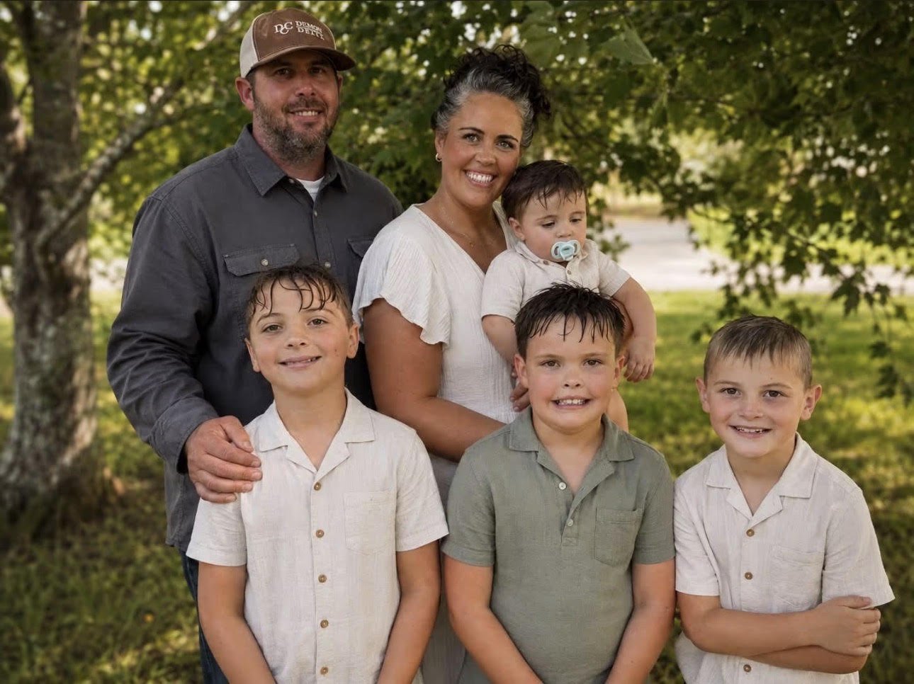 A family of seven, two adults and five children, smiling outdoors with trees and greenery in the background.