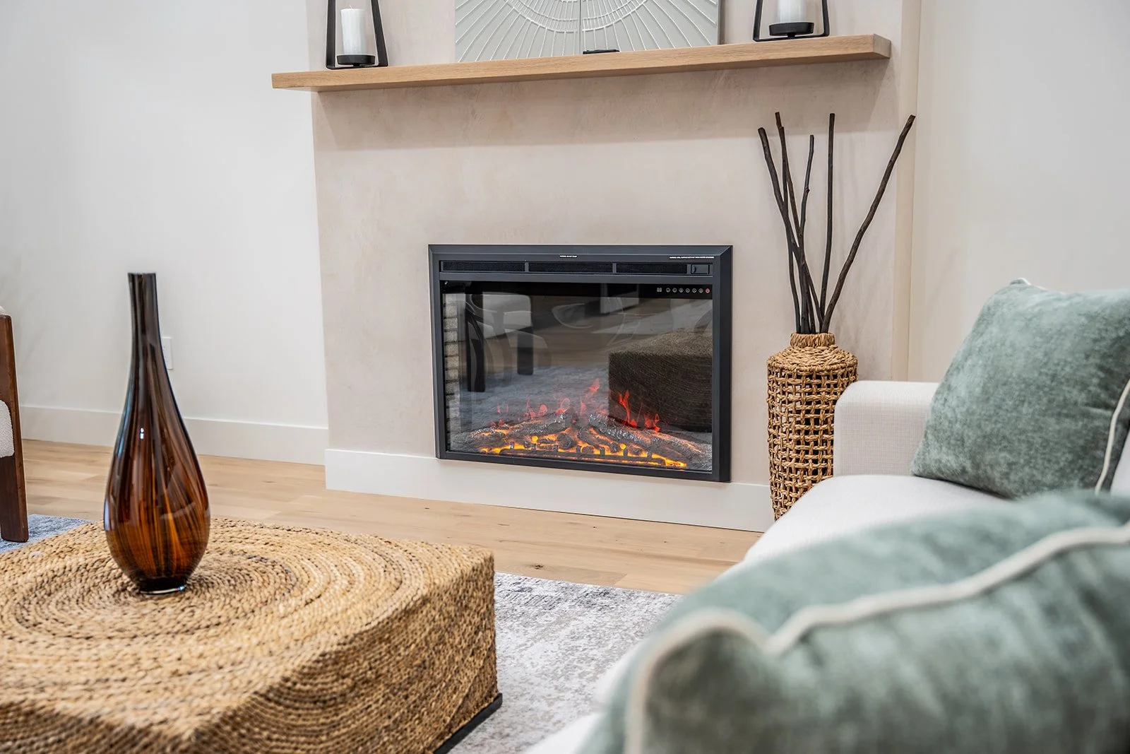 Living room with a modern electric fireplace on a white wall, decorated with a large wicker vase with black sticks, green cushions on a light-colored sofa, a woven circular coffee table with a tall amber glass vase, and a light-colored rug.