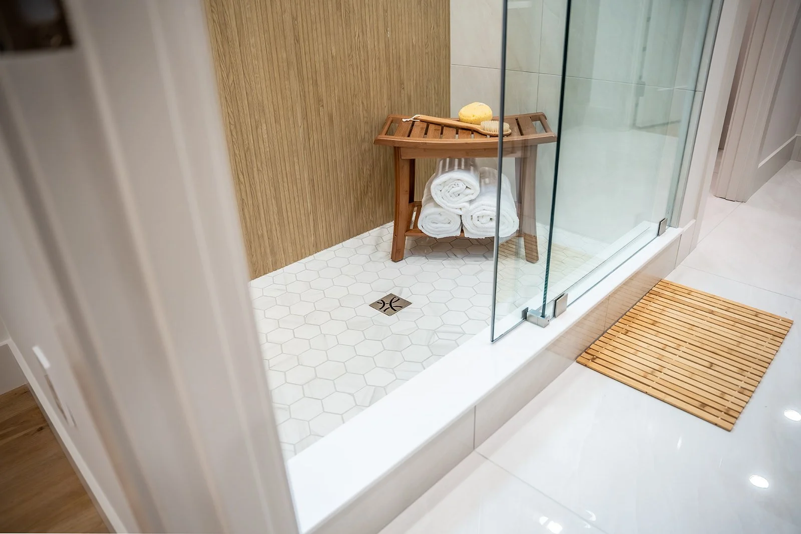 A bathroom shower area with a glass door, wooden wall panel, white hexagonal tiles, and a wooden stool holding rolled white towels, a brush, and a sponge. A wooden mat is outside the shower.