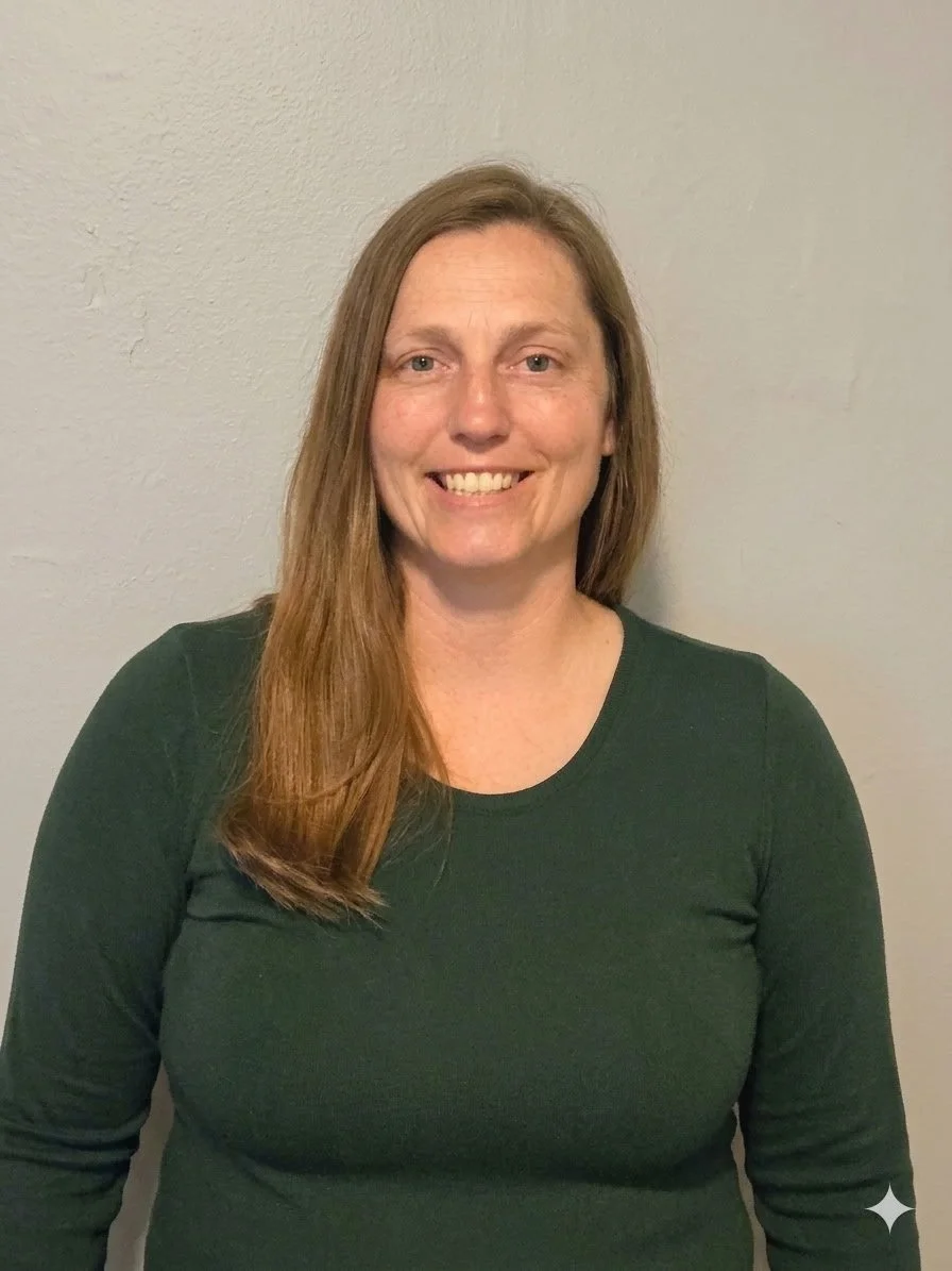 A woman with long brown hair, wearing a dark green long-sleeve shirt, smiling, standing against a plain, light-colored wall.
