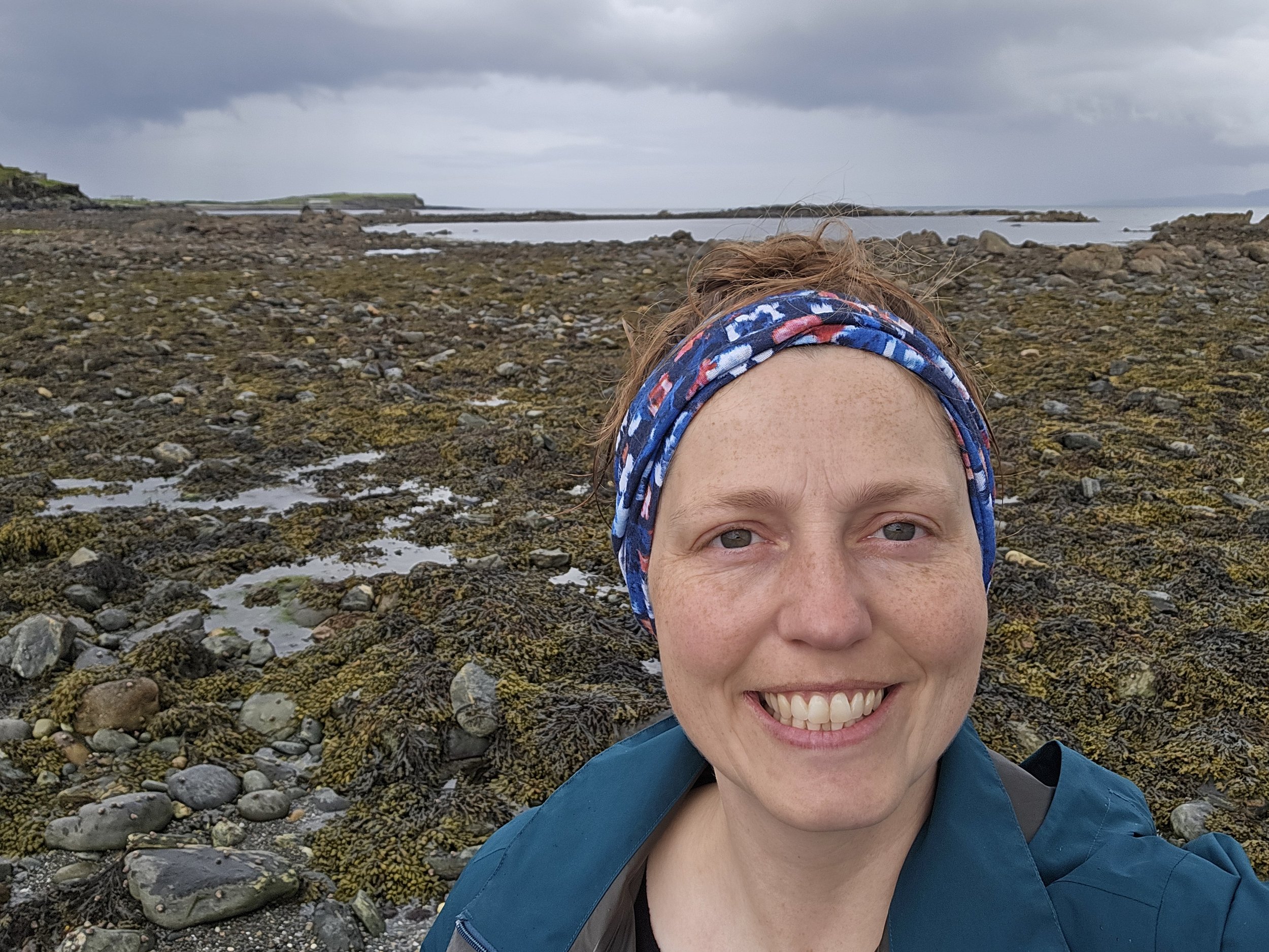 A person smiling at the camera in an outdoor rocky coastal area with cloudy sky, wearing a headband and a blue jacket.