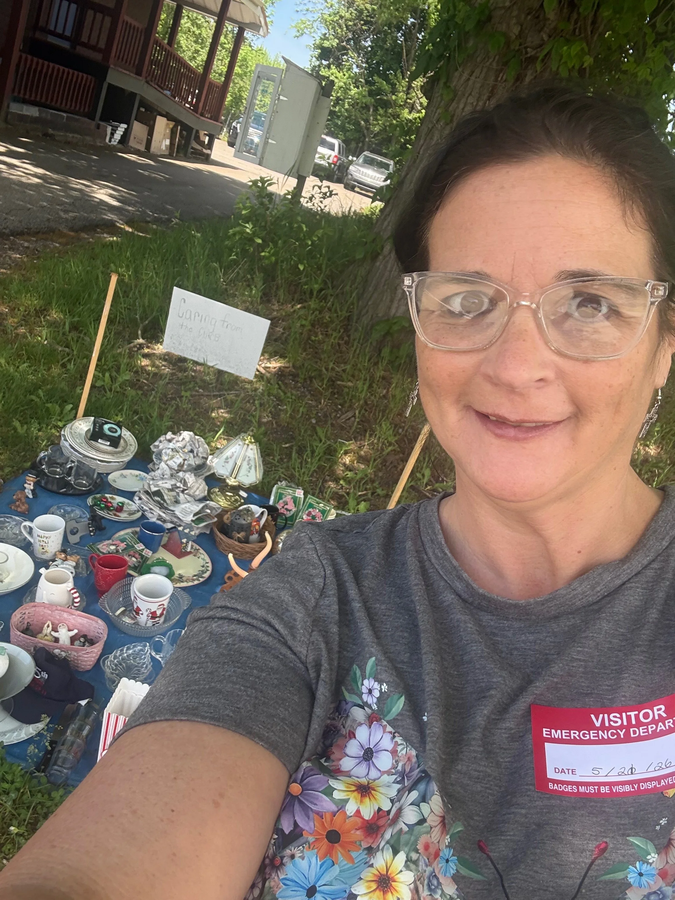 A woman taking a selfie outdoors near a tree and a table of various collectibles and pottery items. She is wearing glasses and a gray shirt with a visitor badge. The scene includes a sign that says 'Caring from the...'